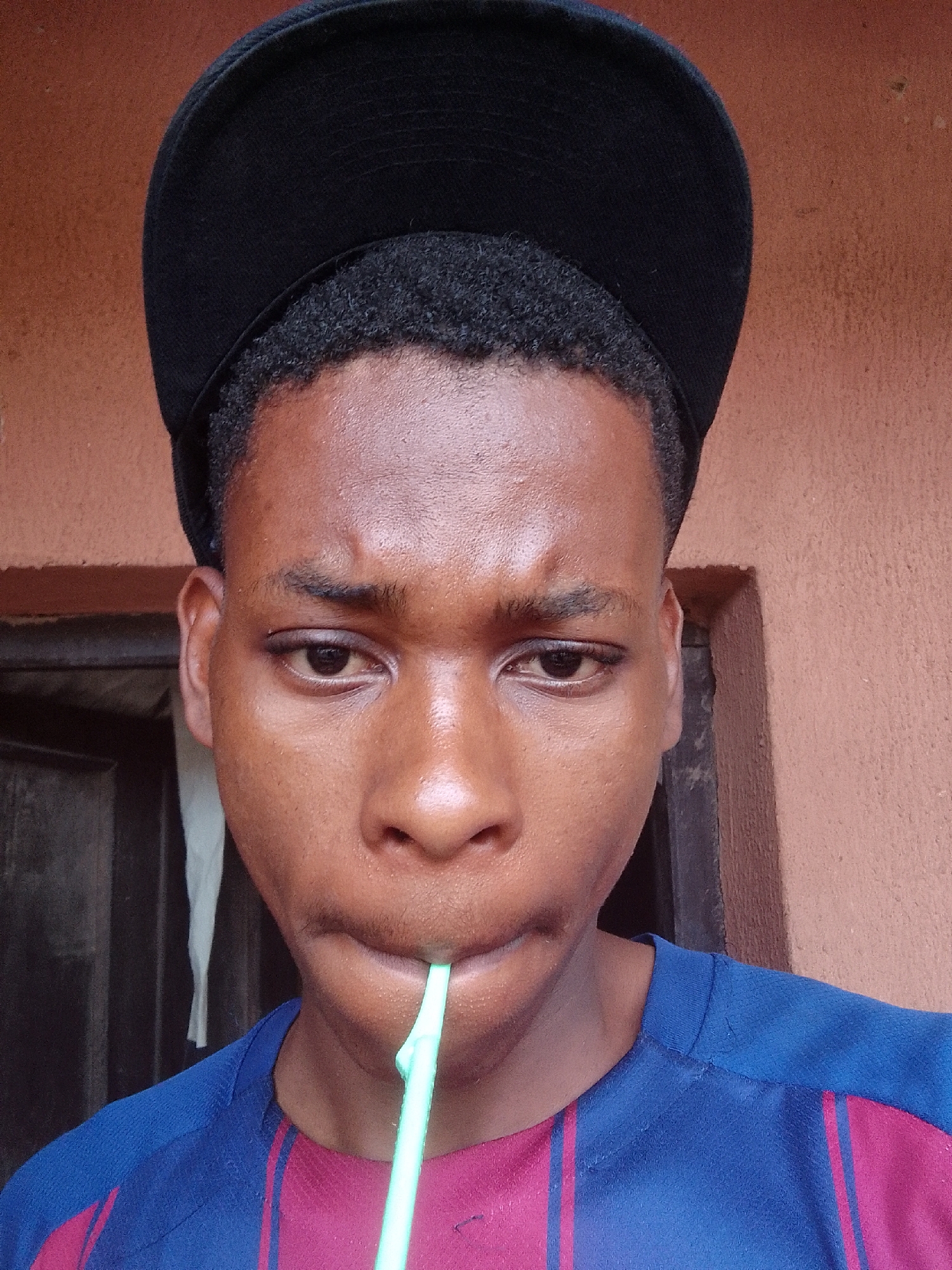 A person with dark skin is drinking from a light blue straw. The person is wearing a black baseball cap and a blue and red shirt. A door with a white curtain is in the background. The person is located in Unknown, Unknown.