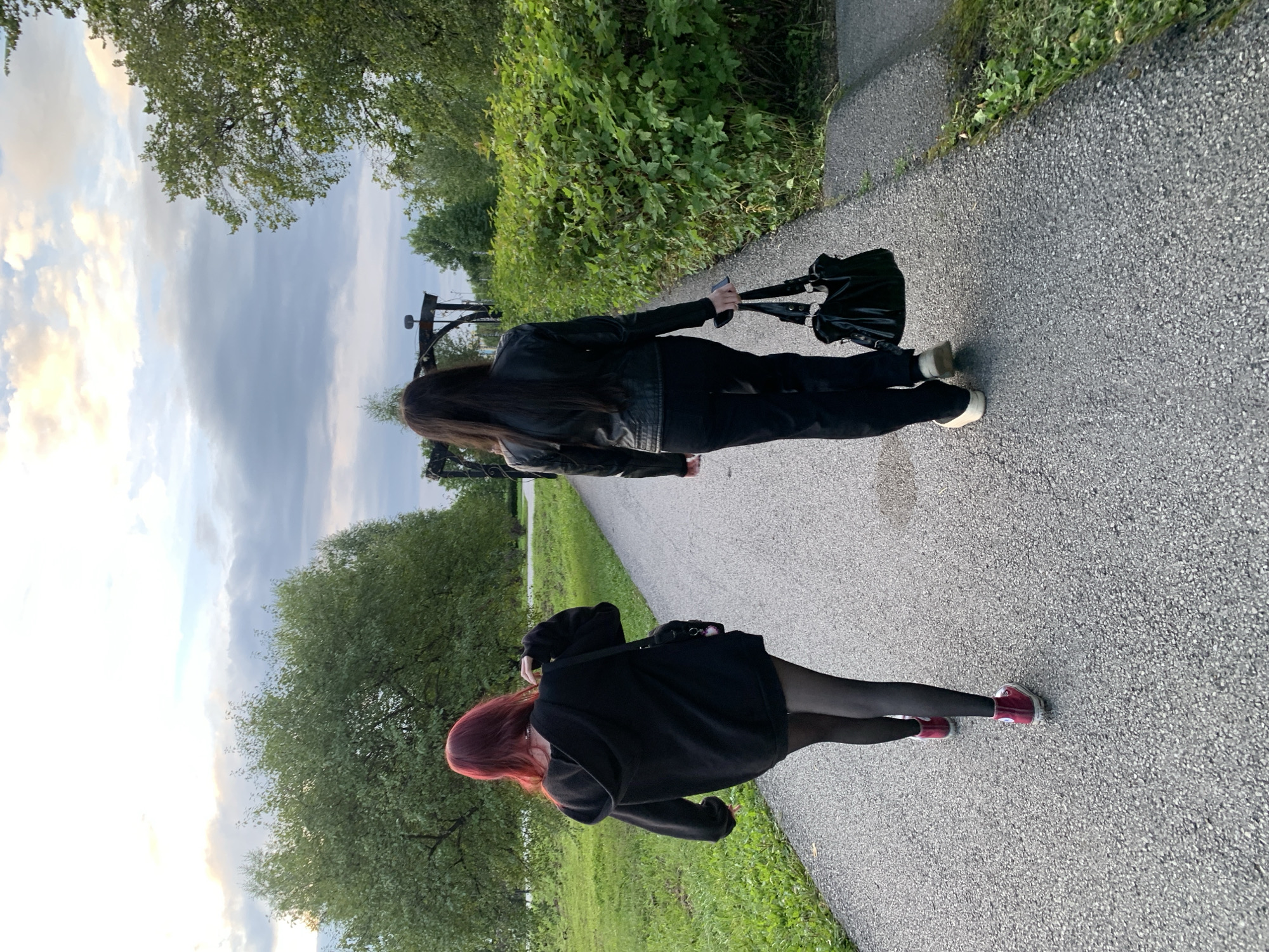 Two individuals are walking away from the camera on a paved path in a park-like setting. The person on the left has red hair and is wearing a black hooded sweatshirt, black tights, and maroon high-top sneakers. They are carrying a black bag over their shoulder. The person on the right has long black hair and is wearing a black leather jacket, black pants, and light-colored sneakers. They are carrying a black handbag. The path is bordered by green grass and dense foliage. In the background, there are trees and a sky with clouds, suggesting it is daytime. A dark, ornate archway structure is visible in the mid-ground. The location is identified as Prokopyevsk, Russia.
