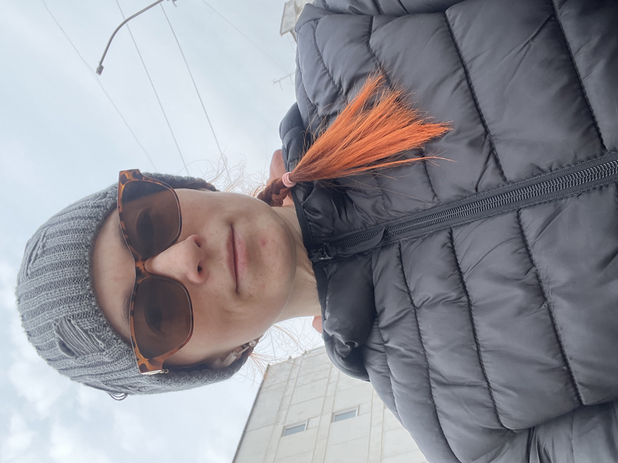 A woman with red hair, sunglasses, and a gray knit cap is seen in an upwards-angled selfie. She is wearing a black puffer jacket. A utility pole is visible behind her and the sky is overcast. A light-colored building is also in view behind her. Based on the presence of the overcast sky and architectural details from the building, it's plausible this photograph was taken in the city of Chaikovsky, Russia.
