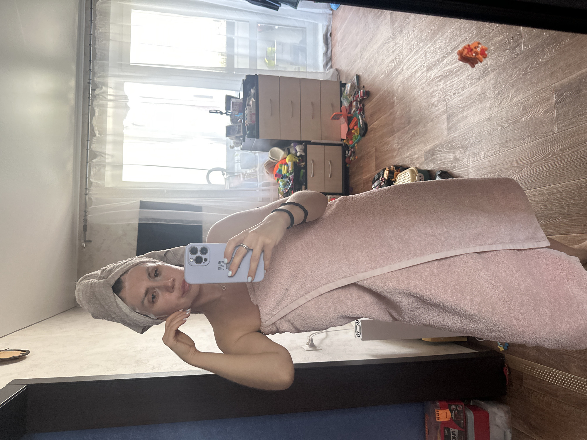 A woman is standing in a room, taking a selfie in a mirror. She has a towel wrapped around her head and another towel wrapped around her body. She is wearing a face mask. The room appears to be a bedroom or bathroom, with a dresser and toys on the floor. The lighting is bright, and the overall scene is casual and domestic. The location is stated to be in Novoaltaysk, Russia, although no specific landmarks or context within the city are provided in the image itself.