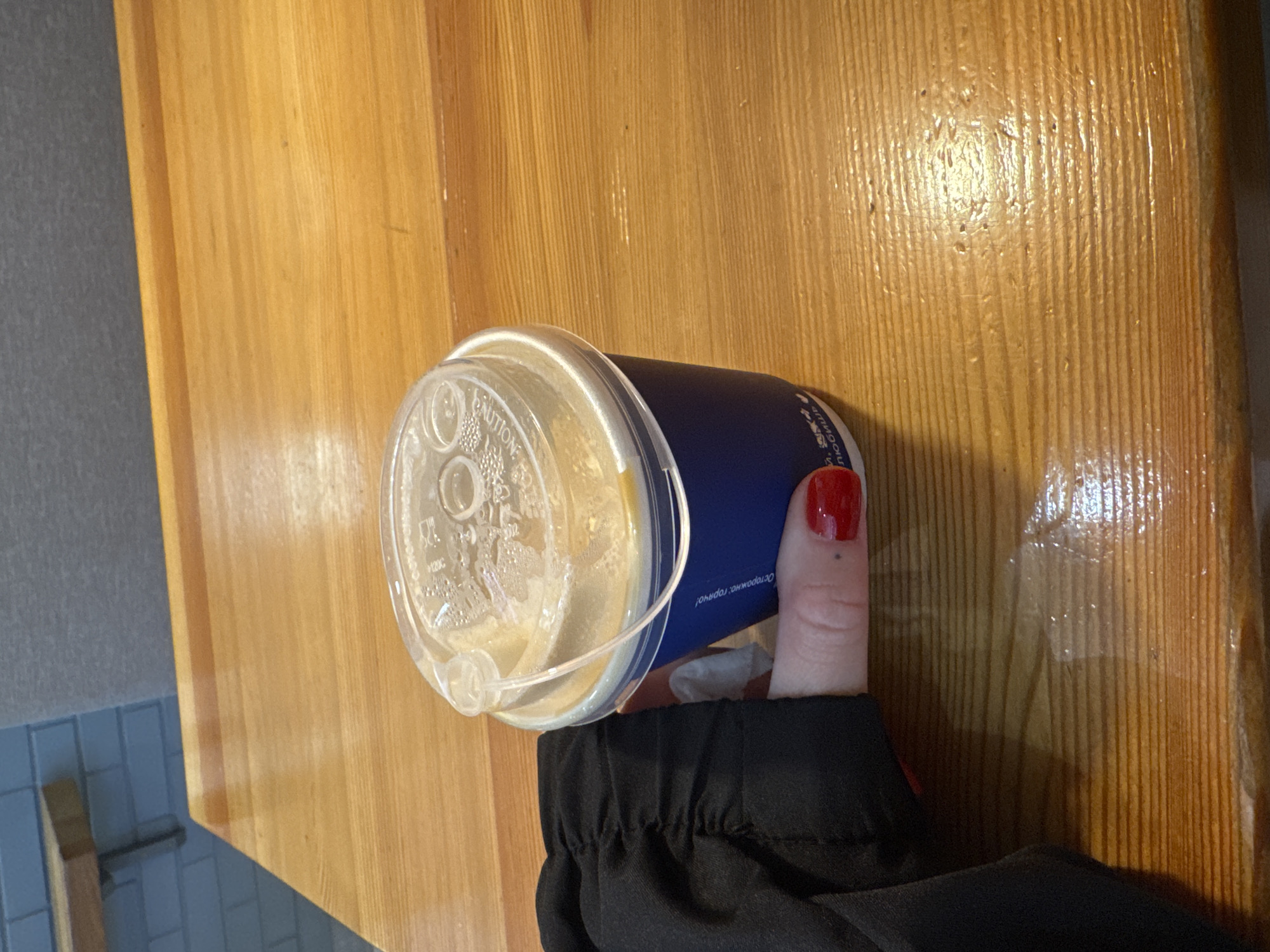 A person holding a blue coffee cup with a clear lid is positioned on a wooden table. The table has a light brown, possibly varnished, wooden surface. The person's thumb with red nail polish is visible holding the cup. The person is wearing a black garment with a gathered sleeve. A glimpse of a blue tiled floor is visible in the lower left corner of the image. The specific location in Rostov-on-Don, Russia, cannot be determined based solely on the visual information in the image.