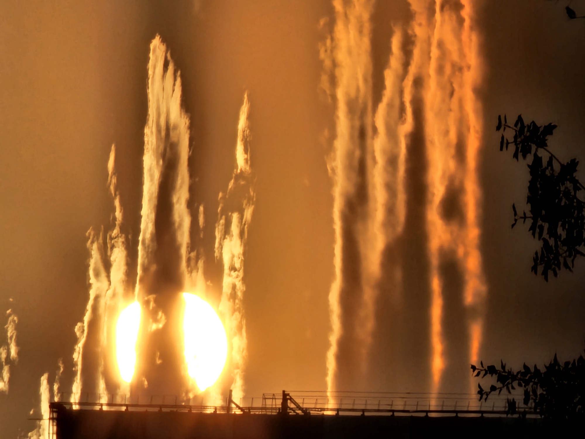 This image captures a dramatic sunset viewed from Khimki, Russia, with the sun partially obscured by clouds and a tall structure resembling a building or industrial facility dominating the left side of the frame. The sky is ablaze with intense orange and yellow hues, indicating the setting sun. Several layers of clouds are visible, some brightly illuminated from behind by the sun, creating a radiant effect, while others are darker, casting shadows. The prominent structure on the left, silhouetted against the bright sky, features scaffolding or external staircases. In the foreground, the dark silhouettes of trees and foliage are visible, adding depth to the scene. The overall impression is one of natural beauty juxtaposed with an urban or industrial landscape.