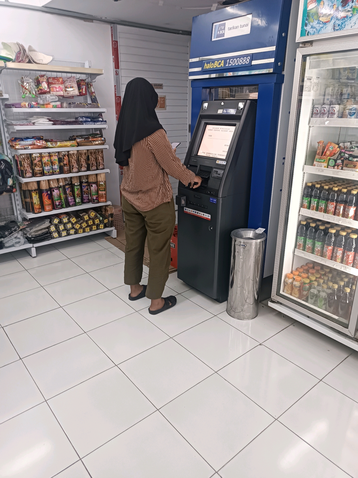 The image depicts an indoor scene, likely a convenience store, in Tabanan, Indonesia. A person wearing a black hijab and a brown and white striped shirt with olive green pants is standing in front of an ATM. The ATM is blue and has "haloBCA 1500888" displayed on it. The person is interacting with the ATM, with their hand on the keypad. To the right of the ATM is a tall, silver trash can. To the far right, a glass-doored refrigerator is filled with bottles of beverages. Shelving units filled with various products, including what appear to be snacks and bottled drinks, are visible to the left of the ATM. The floor is tiled with white square tiles. The overall environment suggests a commercial setting, possibly a minimarket or convenience store, with an ATM machine for customer transactions.