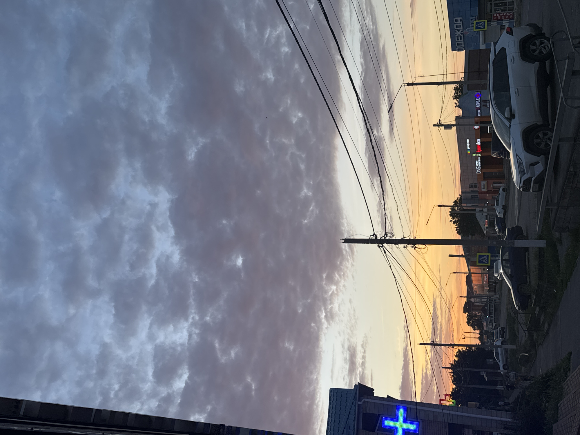 The image depicts a street scene in Yegorlykskaya, Russia, during a sunset. The sky is filled with a mix of dark and light clouds, suggesting a transition in weather or time of day. Utility poles with numerous wires dominate the foreground and mid-ground, stretching across the frame and obscuring parts of the buildings and sky.
Several buildings line the street, some featuring illuminated signs. Visible text includes "ОДЕЖДА" (Clothing) on a blue sign, and other signs with Cyrillic script that are partially obscured or too blurry to read clearly. One sign appears to be for a pharmacy, indicated by a glowing blue cross symbol. A white SUV is parked prominently on the right side of the street, facing away from the viewer, and another smaller, dark vehicle is visible further down the road.
The setting is a typical urban or suburban street with a paved road and sidewalks. The warm hues of the sunset cast a soft glow on the scene, contrasting with the darker clouds. The overall mood is somewhat muted due to the time of day and overcast sky, but the illuminated signs and the sunset provide a sense of activity and visual interest. There are no discernible people in the image. The time of day is clearly sunset, and the weather appears to be partly cloudy.