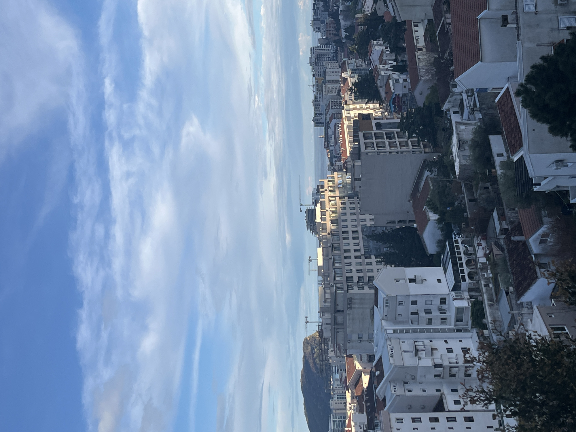 The image displays an elevated panoramic view of the city of Budva, Montenegro, under a partly cloudy blue sky. The Mediterranean Sea is visible on the distant horizon. A large, dark, rocky island or peninsula is prominent on the left side of the frame, jutting out into the sea.
The urban landscape consists of densely packed buildings, ranging from multi-story residential blocks to what appear to be newer, taller commercial or apartment complexes. Architecture varies, with older structures featuring white facades and red-tiled roofs interspersed with more modern, grey-toned buildings with flat roofs. Multiple tower cranes are visible across the cityscape, particularly near the coastline and around newer developments, indicating ongoing construction activity. Patches of green vegetation, including trees and shrubs, are scattered between buildings and properties. The scene is illuminated by natural daylight, casting shadows that suggest either morning or late afternoon. No individuals, specific actions, or interactions are discernible.