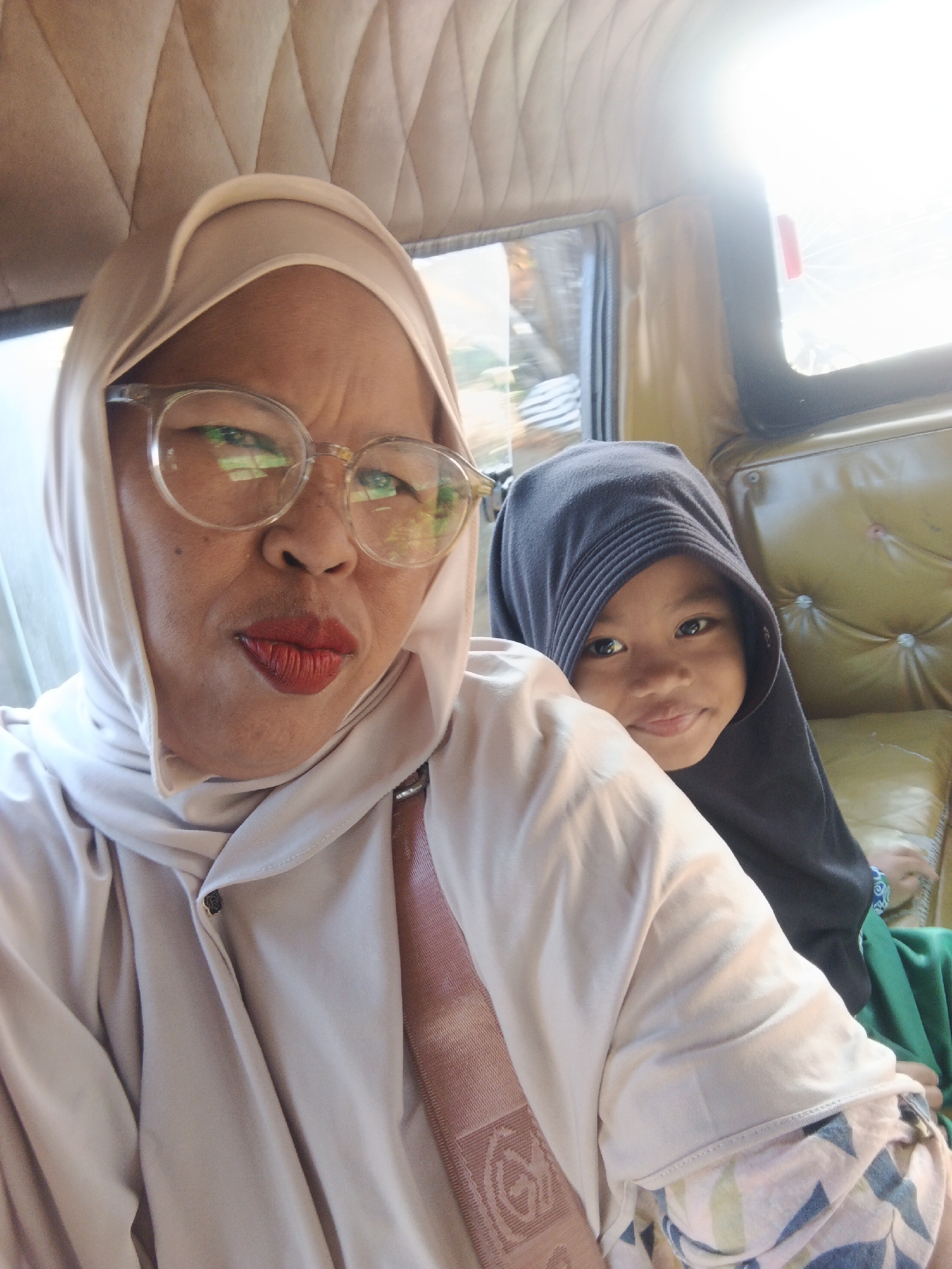 The image shows a selfie taken inside a vehicle, possibly a van or an SUV, with beige upholstered seats and a textured beige ceiling. Two individuals are visible. In the foreground, a woman wearing a light-colored hijab and eyeglasses with clear frames is looking at the camera with her lips pursed and wearing bright red lipstick. She is wearing a light-colored top with a pink strap visible across her chest. Behind her, slightly to the right, a young child wearing a dark hijab is peeking out, looking directly at the camera with a slight smile. The child is wearing a dark hijab and a green garment. The interior of the vehicle appears to be well-lit, with sunlight streaming in from a window on the right, creating a bright glare. The background shows the interior of the vehicle's seating and windows. The location is identified as Warungkiara, Indonesia, suggesting this scene is within that area.