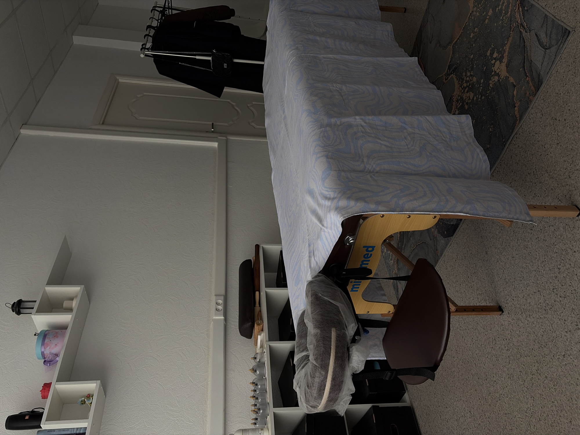 A commercial massage table occupies a room, possibly in Chaikovsky, Russia. The table is covered with a blue and white patterned sheet, and a massage chair is positioned adjacent to it. Shelving units and racks house various objects, including what appears to be professional massage supplies. A decorative wall shelf is visible on the left side of the frame. A dark colored suit jacket hangs on a clothes rack. A gray patterned rug is visible on the floor.
