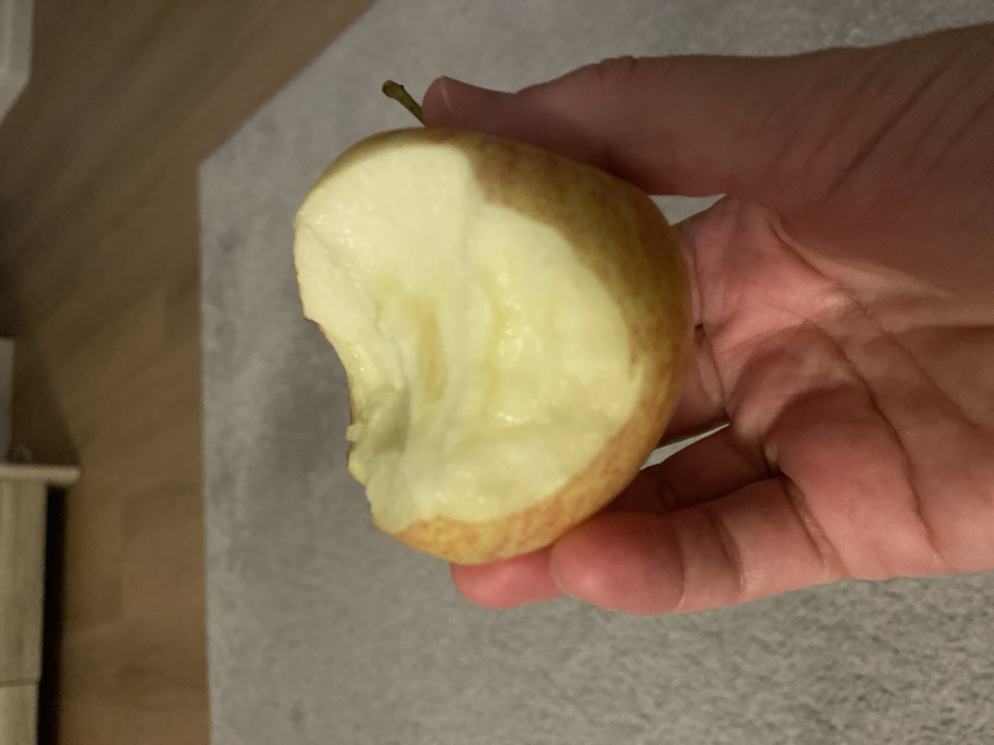 A hand holds a partially eaten apple. The apple is red and yellow, with bite marks revealing its white flesh. The hand is lightly colored and appears to be an adult's. The background is blurred, showing a wooden floor and a grey carpet. The image is taken indoors.