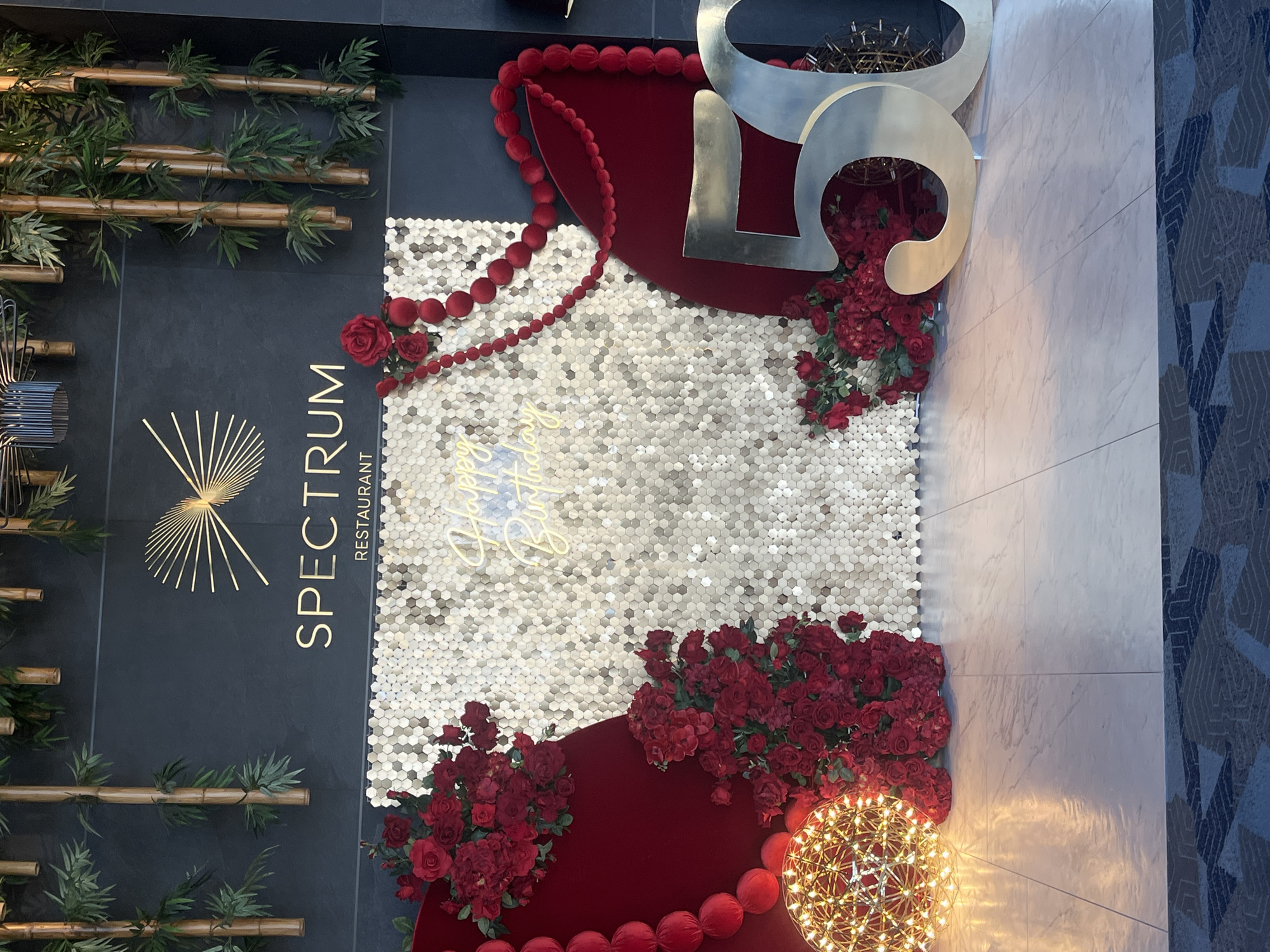 The image shows a wall decoration at the Spectrum Restaurant, likely located in Sofia, Bulgaria, based on the restaurant name. The wall is decorated with a dark gray panel featuring the restaurant's name and logo, and artificial bamboo branches. Adjacent to it is a panel covered in small, hexagonal, silver-colored tiles, adorned with a neon "Happy Birthday" sign. The decoration also includes red fabric elements and strings of red spherical decorations. Red roses are arranged artistically on the panel, and a decorative spherical light fixture is positioned near the bottom. Large silver-colored numbers, seemingly "50", are placed above, with additional rose arrangements. A tiled wall is visible on the right side of the image. The photograph is taken indoors under artificial lighting.