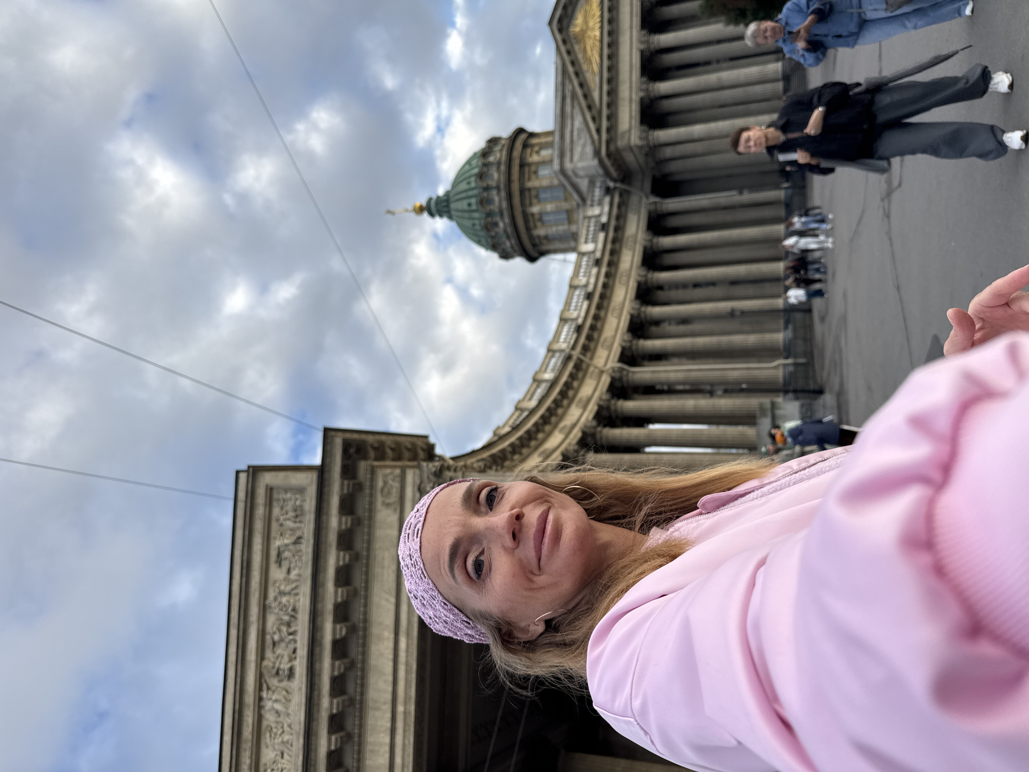 The image shows a woman in a pink jacket and a pink headband taking a selfie in front of the Kazan Cathedral in Saint Petersburg, Russia. The cathedral's iconic colonnade and green dome are visible in the background. Several other people are walking in the square in front of the cathedral. The sky is overcast with clouds.