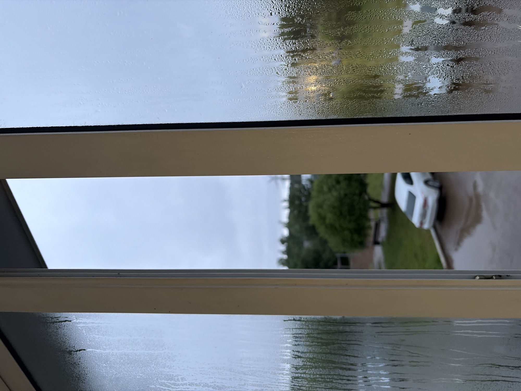 The image depicts a view through a window in Chaikovsky, Russia, on a cloudy, likely rainy day. Condensation streaks are visible on the glass, obscuring the details of the outdoor scene. A white car is parked in a lot, and trees and buildings are discernible in the background. The overall atmosphere suggests overcast weather, and the condensation implies a temperature difference between the interior and exterior environments.