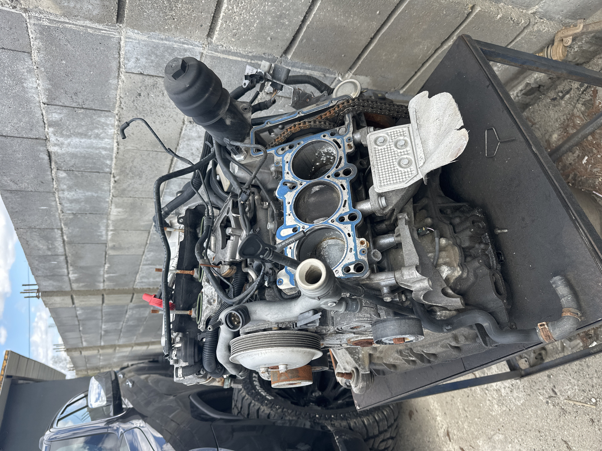 Image depicts a disassembled engine, likely from a vehicle, resting on a dark metal stand. The engine block is exposed, revealing cylinder bores and remnants of a blue gasket material. Numerous hoses, wires, and attached components are visible. The engine is positioned outdoors, near what appears to be a tire, possibly from a vehicle underneath the stand. Above the engine are concrete blocks, suggesting a structure or building nearby. The image was taken outdoors. It is not possible to ascertain the image was taken in Piperevo, Bulgaria. No humans are visible.