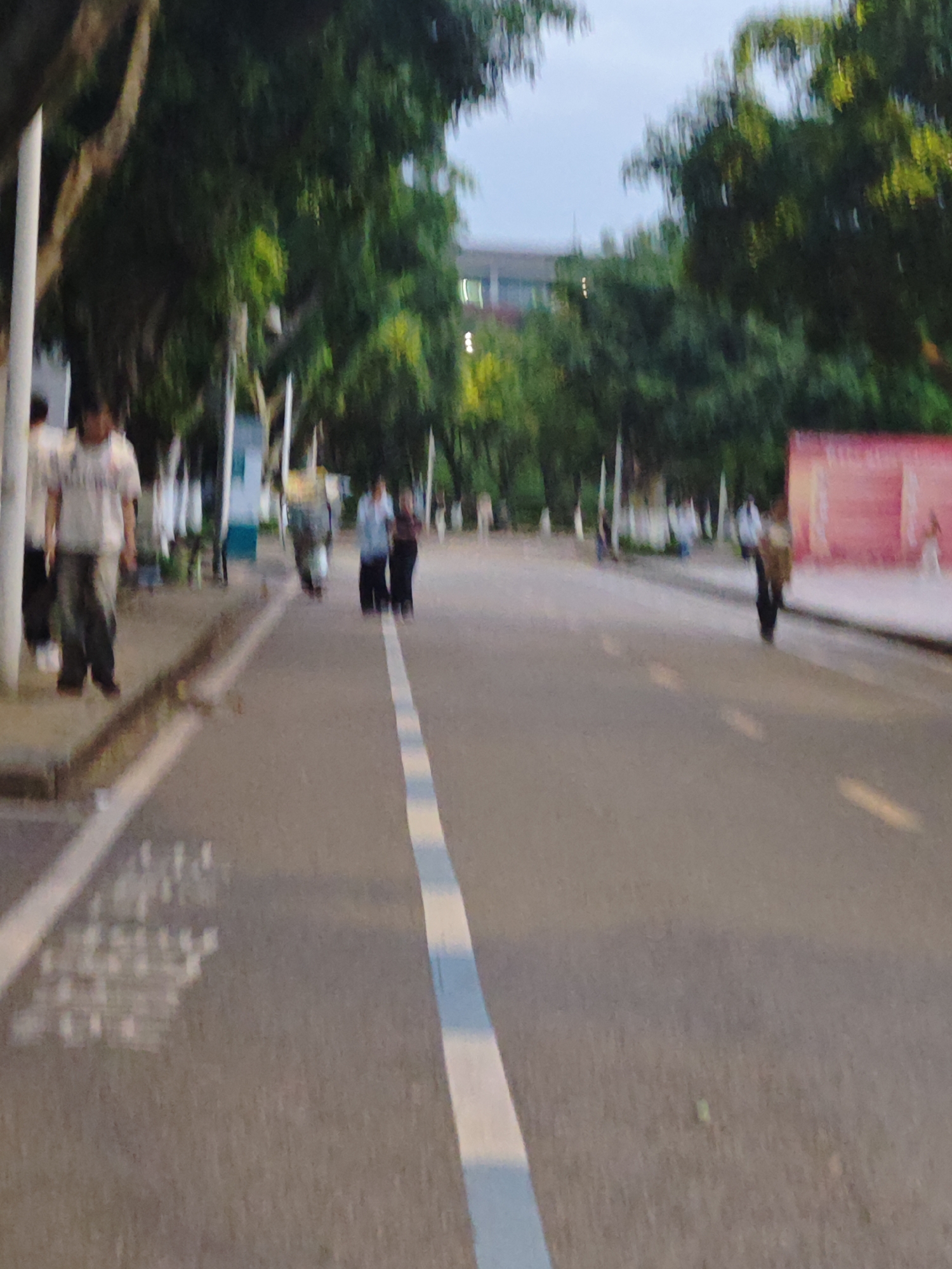 The image shows a blurred street scene in Chongqing High-tech Zone, China. The street has a white line marking the center. Several people are visible, some walking along the sidewalk on the left and others along the street. Trees line both sides of the road. A building is visible in the background. There are white poles along the side of the road and a red structure on the right side.
