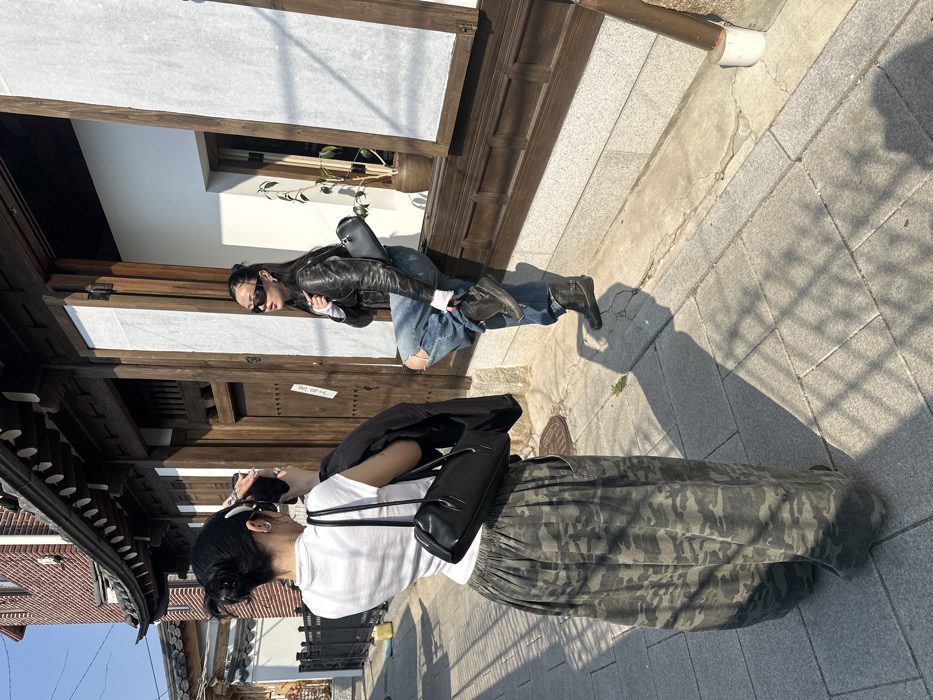 The image captures two individuals on a paved street in the Bukchon Hanok Village, Seoul, South Korea, under direct sunlight. In the foreground on the left, an individual with dark hair styled in a bun, secured by a white clip, faces away from the viewer. This individual wears a white short-sleeved top and green camouflage cargo pants, carrying a black shoulder bag and a black jacket draped over the left arm. They are holding a smartphone with both hands, photographing the second individual. The second individual, positioned on the right, wears dark sunglasses, a black leather jacket over a white long-sleeved shirt, ripped blue jeans, and dark boots. This individual poses with a bent left leg, foot elevated onto a wooden window sill, and leans against the dark wooden frame of a traditional Hanok building's window, which features white paper panels. A small black shoulder bag is worn over their right shoulder. The building's facade includes a granite foundation, dark wooden frames, and visible traditional design elements. A potted plant is visible through the window behind the posing individual. Strong shadows are cast on the light-colored, stone-paved ground. A red brick building is partially visible in the background to the left.