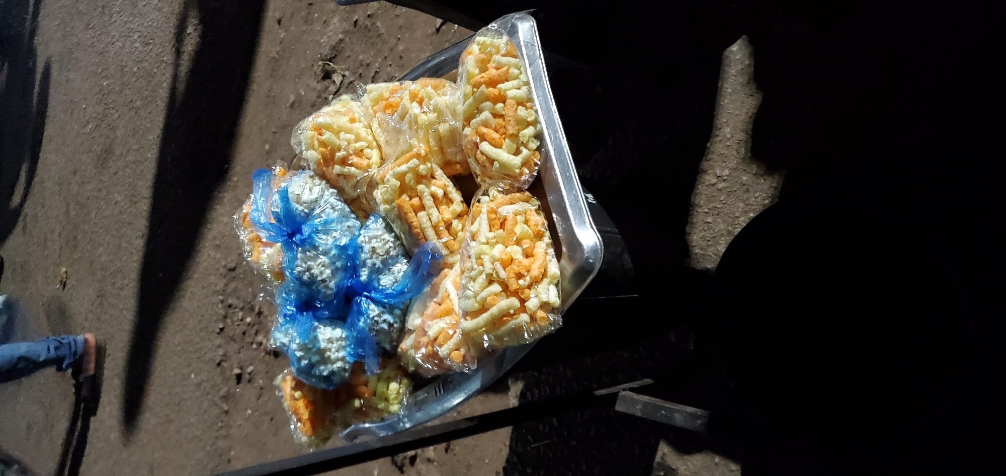 A high-angle shot shows an aluminum tray filled with numerous plastic bags of snacks. Some bags contain yellow and orange cylindrical snacks, while others contain white popcorn-like snacks packaged in blue plastic. The tray rests on the ground or a low surface outside, as indicated by the dirt and pavement surrounding it. To the lower left, a person's feet, clad in jeans, are partially visible. The right side of the image fades into deep shadow. The location appears to be an outdoor urban environment, potentially a street or market in the city of Unknown, Unknown.