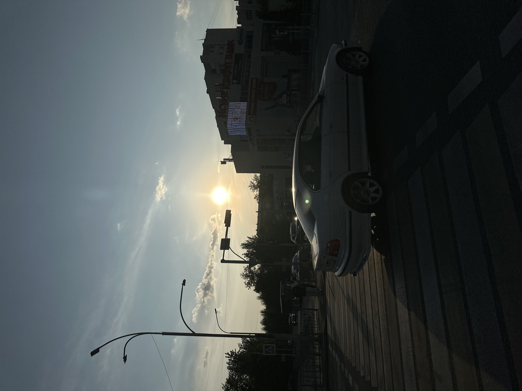This is a photograph taken in Kokshetau, Kazakhstan, at sunset. The image is oriented vertically, with the sky and sun occupying the left side and a street scene on the right.
The sun is low in the sky, casting a warm, golden light that illuminates some of the clouds. The sky is a pale blue with wispy clouds. In the distance, there is a multi-story building with various signs. One prominent sign reads "HOME CREDIT BANK" in large letters. Other text visible on the building includes "Адвокат И ПАРТНЁРЫ" (Lawyer and Partners) and what appears to be a phone number and advertising for a deposit.
A light-colored car is parked on the right side of the image, facing away from the viewer. The car is partially silhouetted by the setting sun. The car is positioned on a pedestrian crossing, indicated by the striped pattern on the road. Streetlights with curved arms are also visible, some illuminated and others not. Trees are present in the background, adding to the urban landscape.
The overall mood is calm and serene, typical of a sunset. The presence of the car and pedestrian crossing suggests an urban environment. The time of day is clearly evening, due to the low angle of the sun and the long shadows cast on the road.