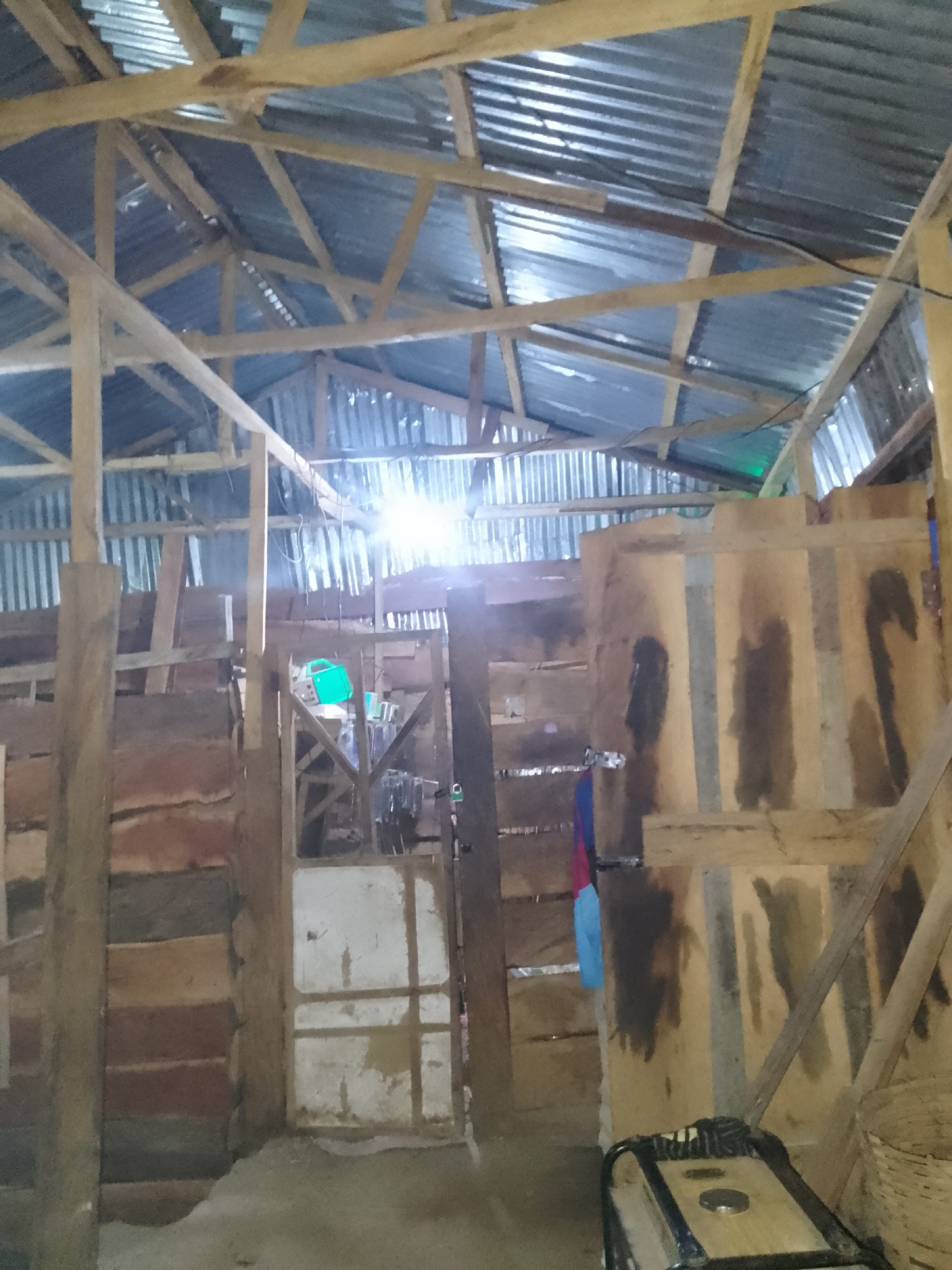 The image depicts the interior of a dimly lit, rustic structure, possibly a workshop or storage area, in Serti, Nigeria. The roof is constructed of corrugated metal sheets supported by a wooden framework. The walls are a patchwork of rough-hewn lumber and corrugated metal. In the center of the image, a doorway leads to an inner area where a bright light source is visible, likely a light bulb. To the left of the doorway, stacks of what appear to be large pieces of lumber are stored. To the right, a portable generator is visible in the foreground. The floor is covered with dirt and debris. There are no people visible in the scene. The overall impression is one of a functional, yet unfinished or unkempt, space.