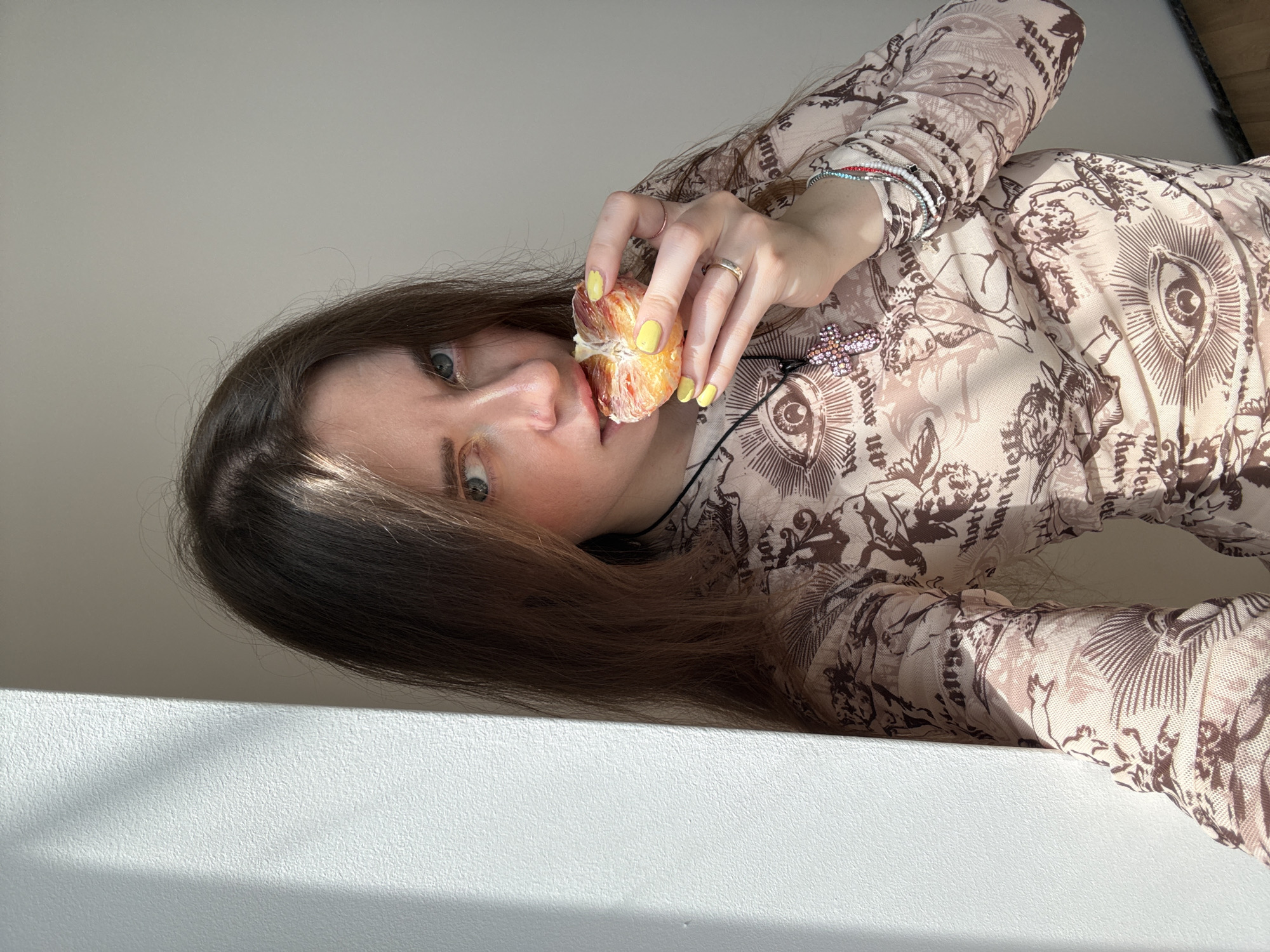 A woman is shown in a selfie while standing next to a white wall. The selfie appears to have been taken from a low angle. The woman has long brown hair that partially covers her face. She is wearing a patterned long-sleeved shirt with a print of eyes, angels, and text. The shirt is cream-colored with brown designs. She is holding a segmented citrus fruit, which appears to be an orange, close to her mouth. Her fingernails are painted yellow. She is wearing a ring on her ring finger and multiple bracelets on her wrist. She is also wearing a necklace with a pink and white cross pendant. The background consists of a plain white wall. The location in the city of Moscow, Russia cannot be determined from the image.