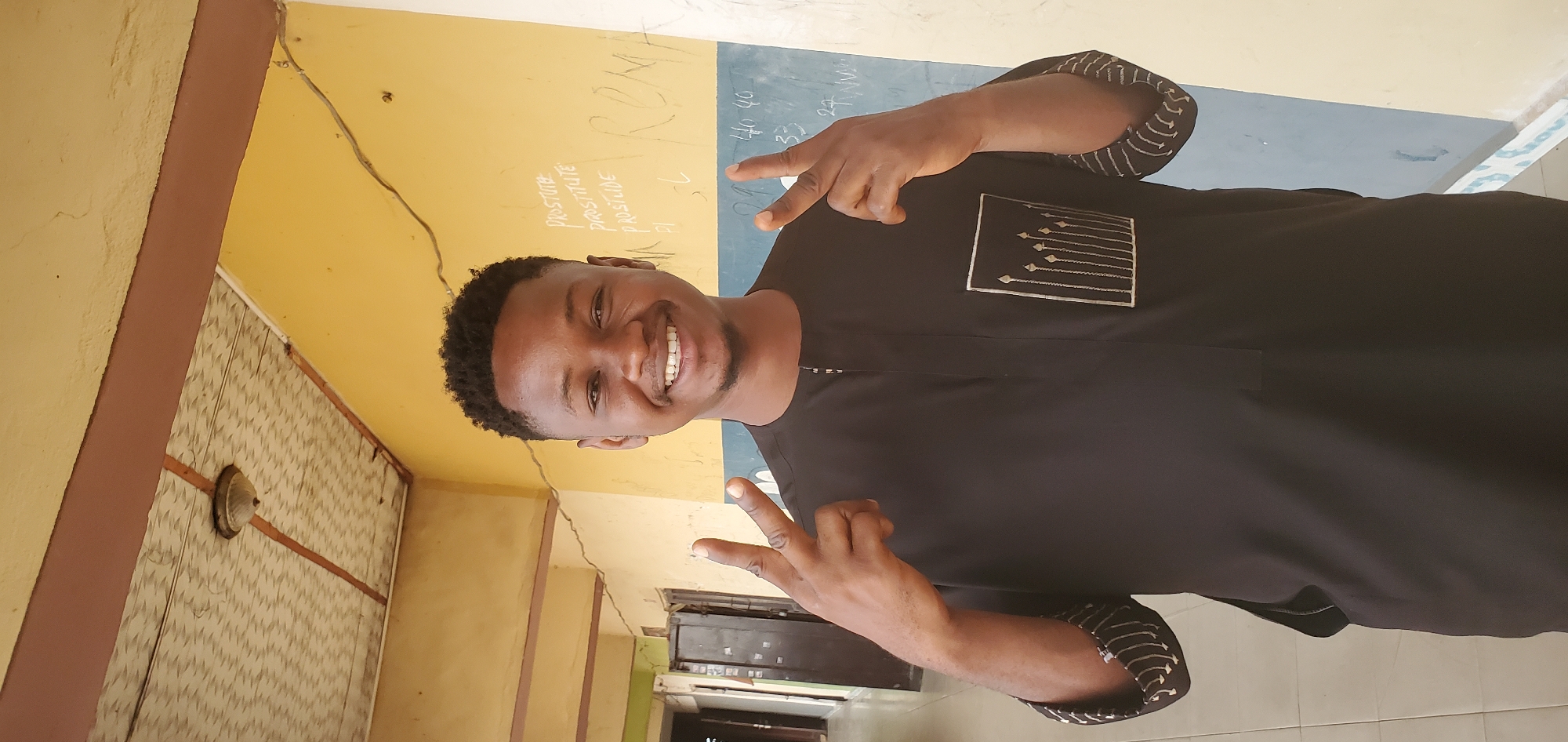 A young man is standing in a room with painted yellow walls and a blue-topped wall with writing. The man is wearing a black outfit with silver stitching around the pocket and the cuffs. He is smiling and making a peace sign with both hands. An electrical outlet is visible on one of the walls. The location is Mararaba, Nigeria.