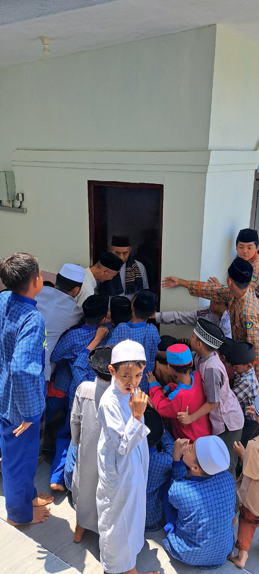 In Sumber Gedang, Indonesia, a group of Muslim children, some wearing peci caps and traditional Islamic clothing, gather around a doorway. Two older men, also wearing peci caps and traditional attire, are entering or exiting the building through the doorway. One man wears a striped scarf. The building appears to be a mosque or similar religious structure, with a white exterior wall and a simple doorframe. The children are observing the men, with some reaching out towards them. The lighting suggests it is daytime, with sunlight casting shadows on the ground. The scene conveys a sense of community and respect within the religious gathering.