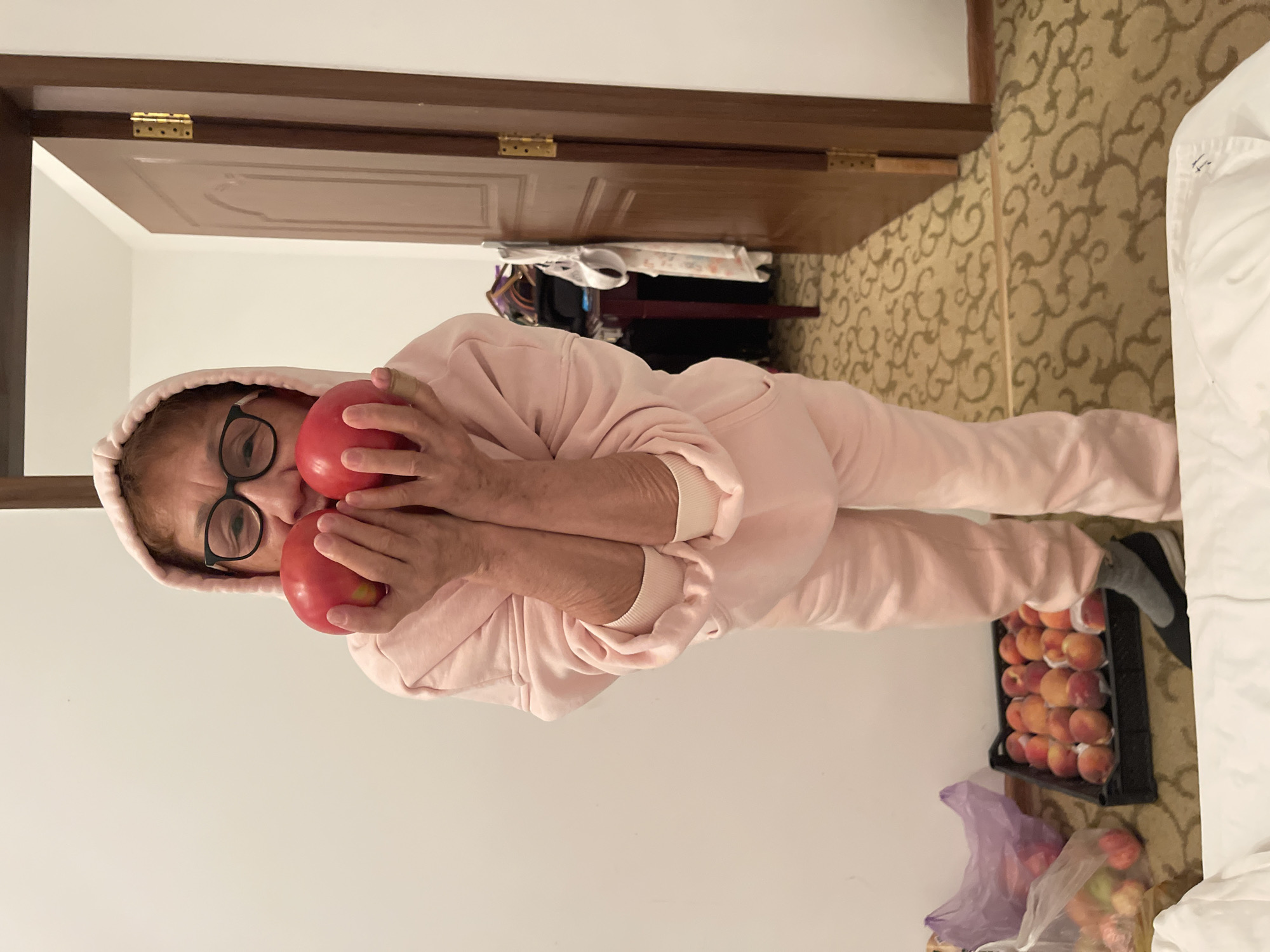 An older woman wearing a pink hoodie and pants, with glasses, is holding two red apples to her face. In the background, a doorway to a room is visible, and there is a crate of peaches on the floor, along with plastic bags of fruit. The scene appears to be indoors, possibly in a hotel room or apartment, in the city of Ornek, Kyrgyzstan.