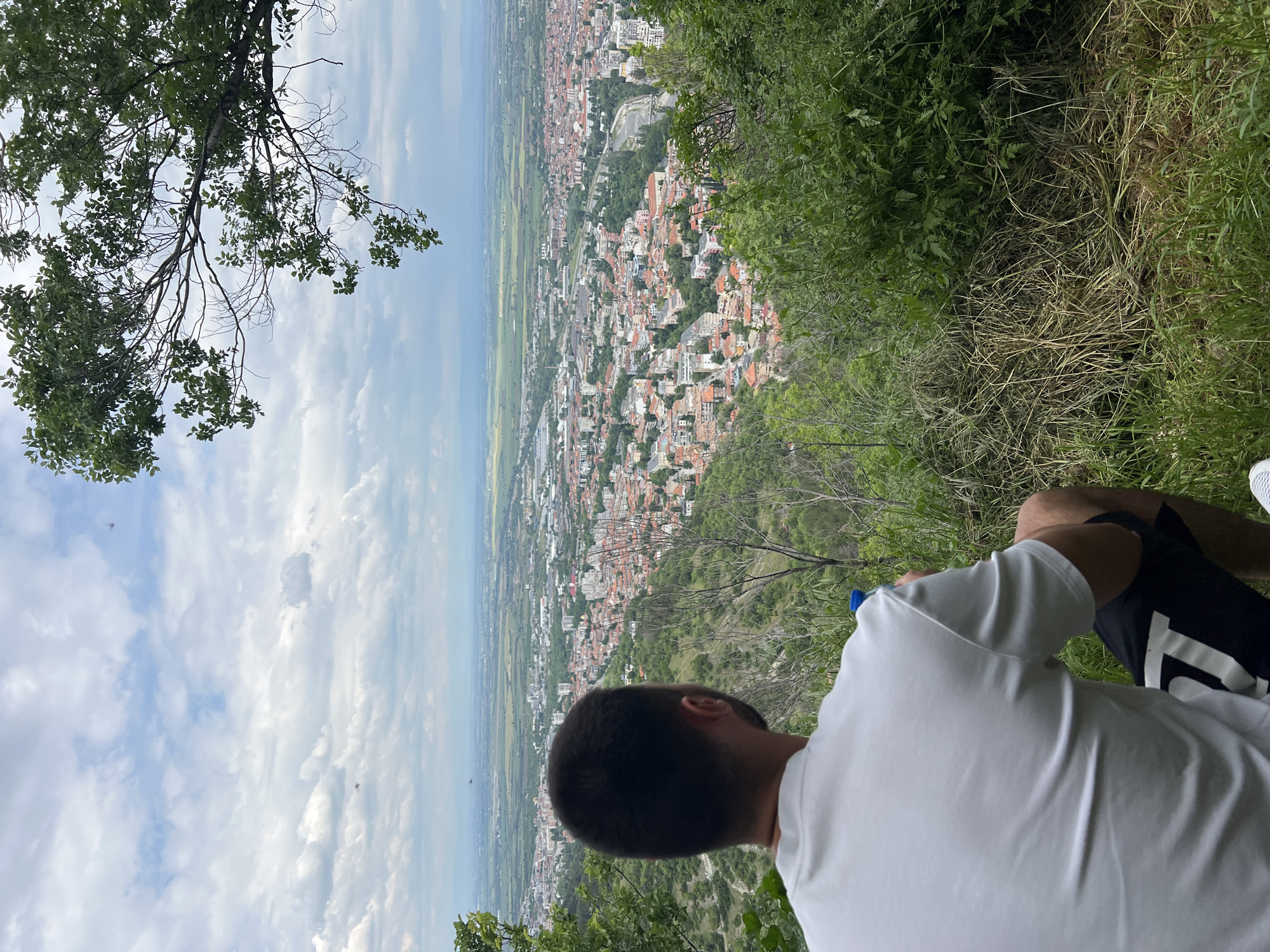 A view from above Asenovgrad, Bulgaria. A person sits on an incline covered in dense vegetation, looking over the city. The foreground includes a branch of a tree on the left and the man's hair, white shirt, and a glimpse of dark shorts with a white design on the right. The city stretches out in the mid-ground with a mix of buildings and green spaces. In the background, there is a broad expanse of agricultural land and the sky, with its scattered clouds, fills the upper portion of the frame.