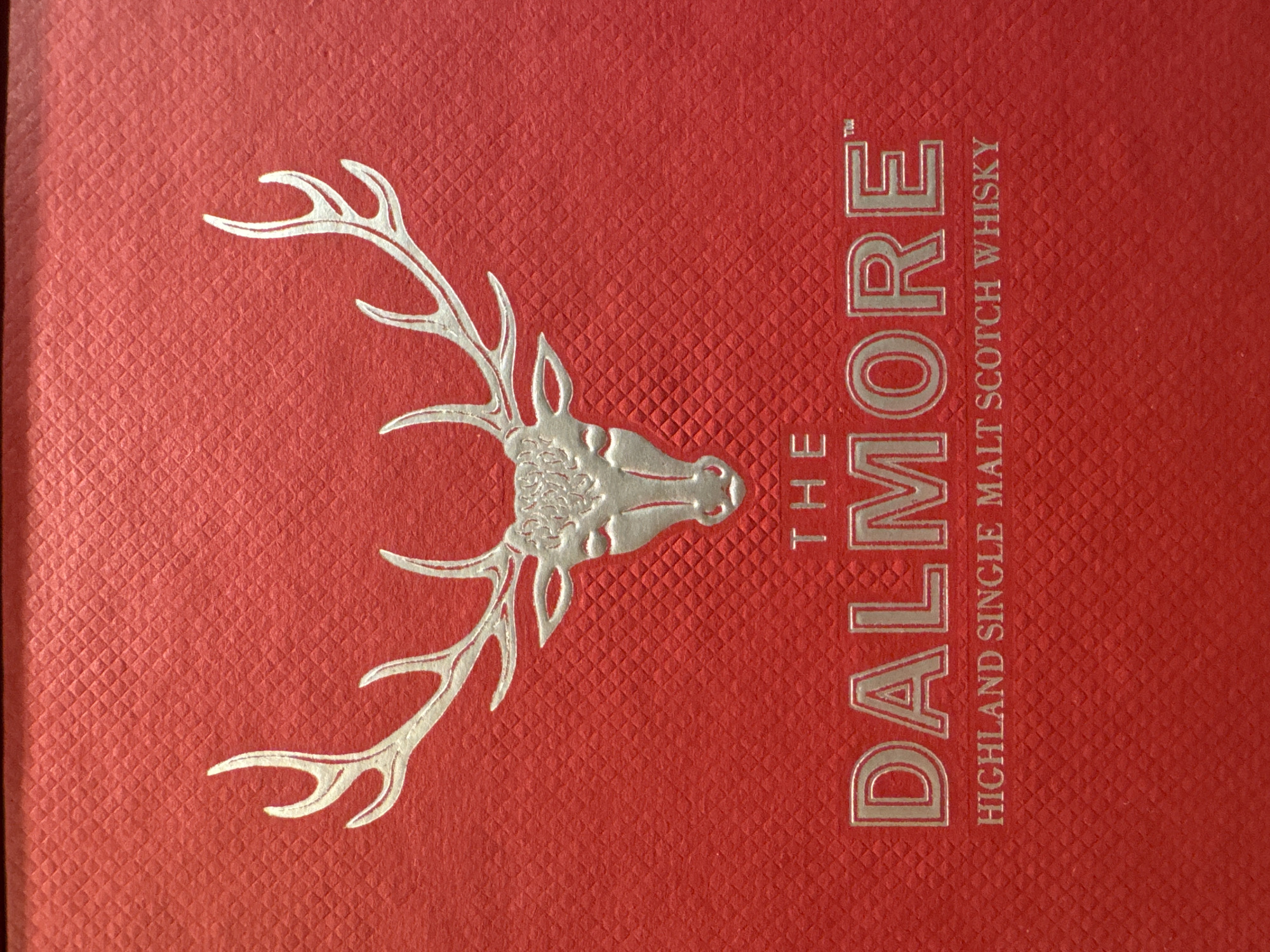 The image shows a close-up of the front of a Dalmore Highland Single Malt Scotch Whisky box. The box is red with a textured surface. A silver stag head with antlers is located on the left side of the box. To the right of the stag head is the text "THE DALMORE" in silver, vertically aligned. Below "THE DALMORE" and oriented horizontally is the text "HIGHLAND SINGLE MALT SCOTCH WHISKY". The image does not provide any information about the location of Chaikovsky, Russia.