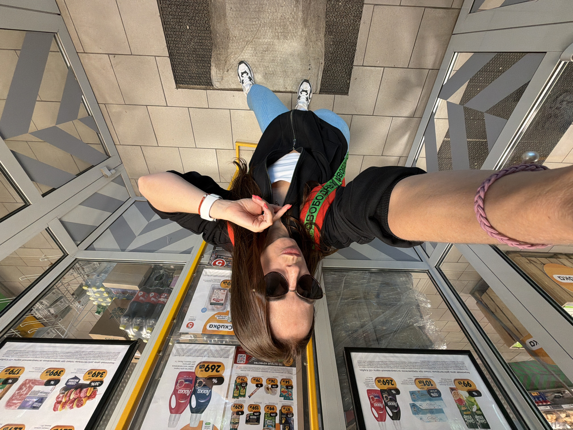 An overhead selfie captures a young woman standing on a light-tiled floor with dark grey matting, likely inside a supermarket in Birobidzhan, Russia. The perspective makes her appear inverted in the frame, with her head towards the bottom and feet towards the top.
The woman has long brown hair and wears dark sunglasses, a black zippered jacket over a white top, light blue jeans, and black and white sneakers. A green and red strap with Russian text, possibly from a bag or garment, crosses her chest. She wears a red bracelet and a white smartwatch on her left wrist, and a pink braided bracelet on her right. Her right hand is raised near her face, making a gesture.
The setting is clearly a retail environment, specifically a grocery store or supermarket. Large glass-topped display cases line the area, showcasing various products and promotional materials. Visible Russian text on the signs includes "АКЦИОННЫЕ ЦЕНЫ ДЕЙСТВИТЕЛЬНЫ ТОЛЬКО ПРИ ПРЕДЪЯВЛЕНИИ АКТИВИРОВАННОЙ КАРТЫ «Х5 КЛУБА»" (Promotional prices are valid only upon presentation of an activated "X5 Club" card), indicating an X5 Retail Group store. Other discernible text includes brand names like "Ласка" (Laska) for laundry detergent, "Global Village Vintage," and various prices in rubles, such as 99, 101, 159, 269, 349, 399, and 179. The interior is brightly lit, suggesting daytime.