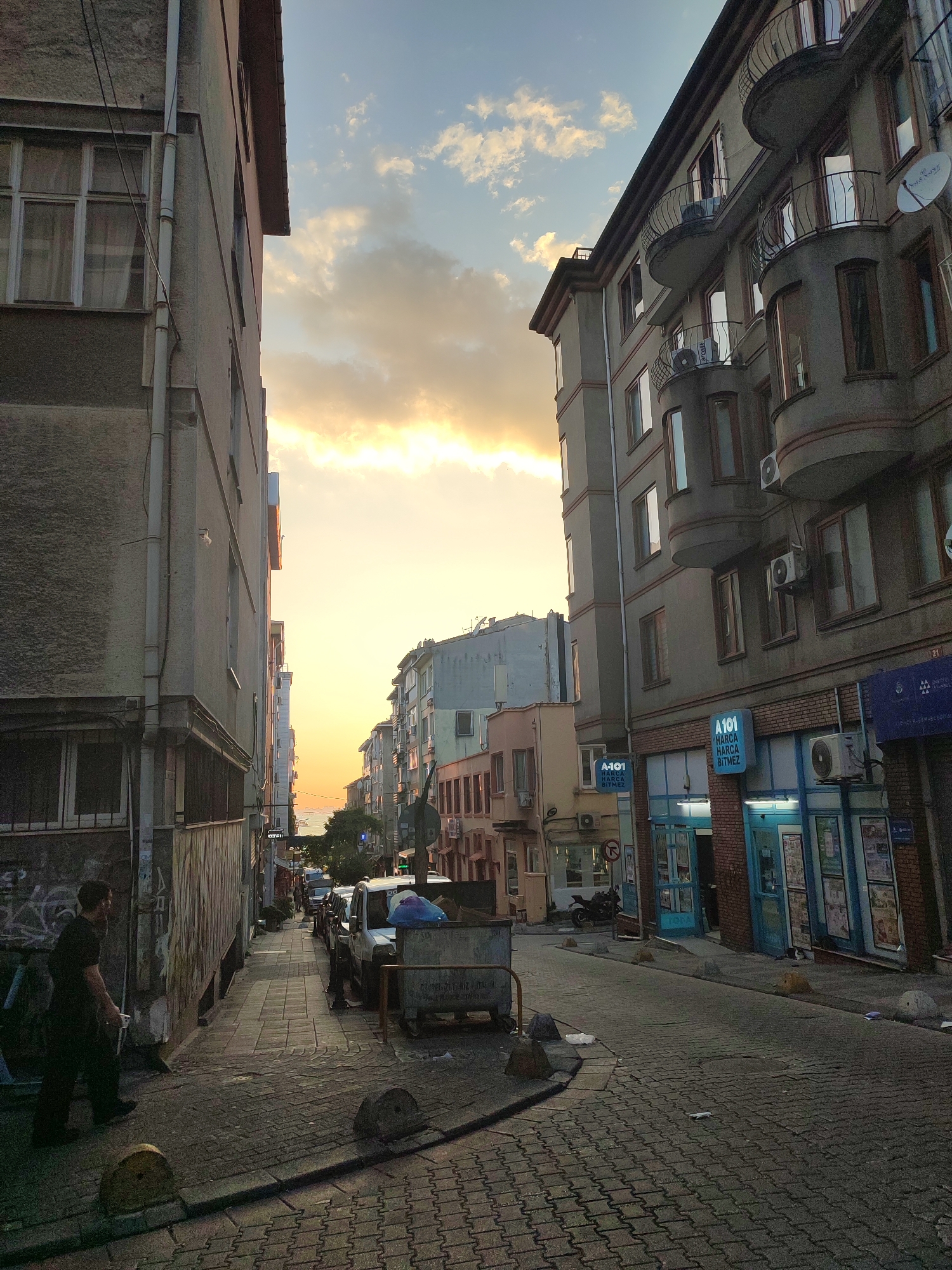 The image depicts a narrow city street in Kadıköy, Turkey, bathed in the warm glow of a sunset. The street is paved with interlocking bricks and flanked by multi-story buildings on both sides. The building on the left is a simple, weathered structure with visible pipes and windows. The building on the right is more ornate, featuring curved balconies with wrought-iron railings and air conditioning units. Several shops with blue signage are visible on the ground floor of the building on the right, one of which reads "A101".
A lone man is walking down the sidewalk on the left side of the street, carrying a bag. Parked cars and a dumpster are visible further down the street. The sky above is filled with soft, scattered clouds, illuminated by the setting sun, creating a dramatic effect of light and shadow across the urban landscape. The overall scene conveys a sense of everyday life in a Turkish city at dusk.
