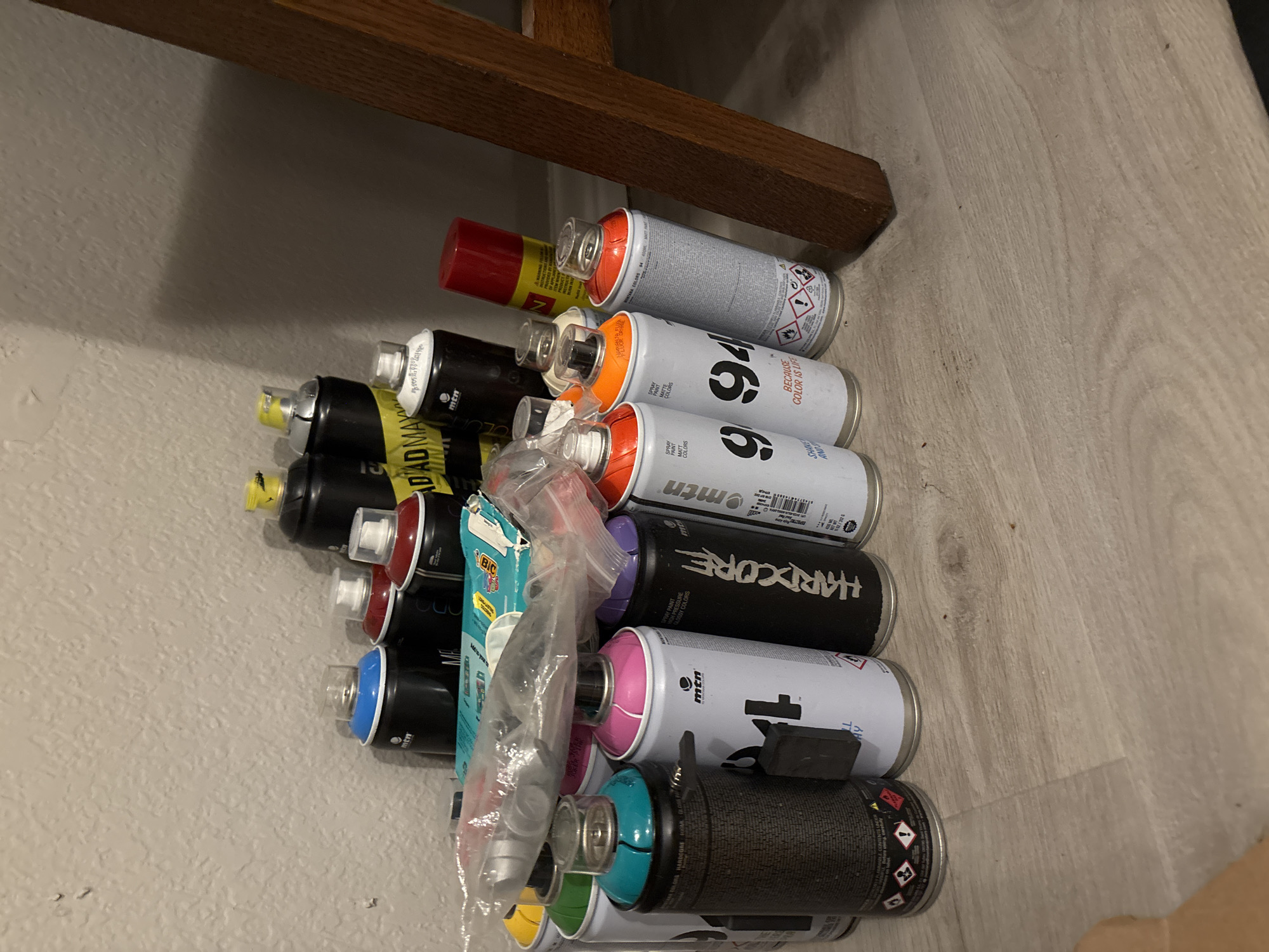 A collection of spray paint cans is arranged on a light-colored, textured floor against a textured, light-colored wall. The cans are of various colors and brands, including MTN, Hardcore, and others. Some cans are upright, while others are tilted. A clear plastic bag containing what appear to be can caps is draped over some of the cans. A portion of a wooden chair leg is visible on the right side of the image. The scene appears to be indoors, possibly in a storage area or workspace.