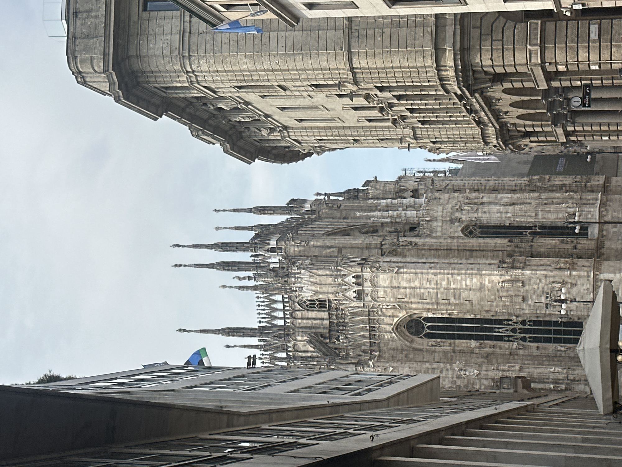 The image captures a street view in Milan, Italy, dominated by the magnificent Duomo di Milano, a gothic cathedral, and flanked by two imposing buildings. The Duomo's intricate spires and stonework are visible, bathed in soft, overcast light. To the left, a modern building with a minimalist facade and numerous windows lines the street. To the right, a more ornate, older building with classical architectural elements, including arched windows and decorative sculptures, stands prominently. Two flags, one with horizontal blue and green stripes and a white element, and another blue flag with a white diagonal cross, are visible against the sky. The street appears to be relatively empty, with only hints of activity such as streetlights and a canopy suggesting an outdoor seating area.