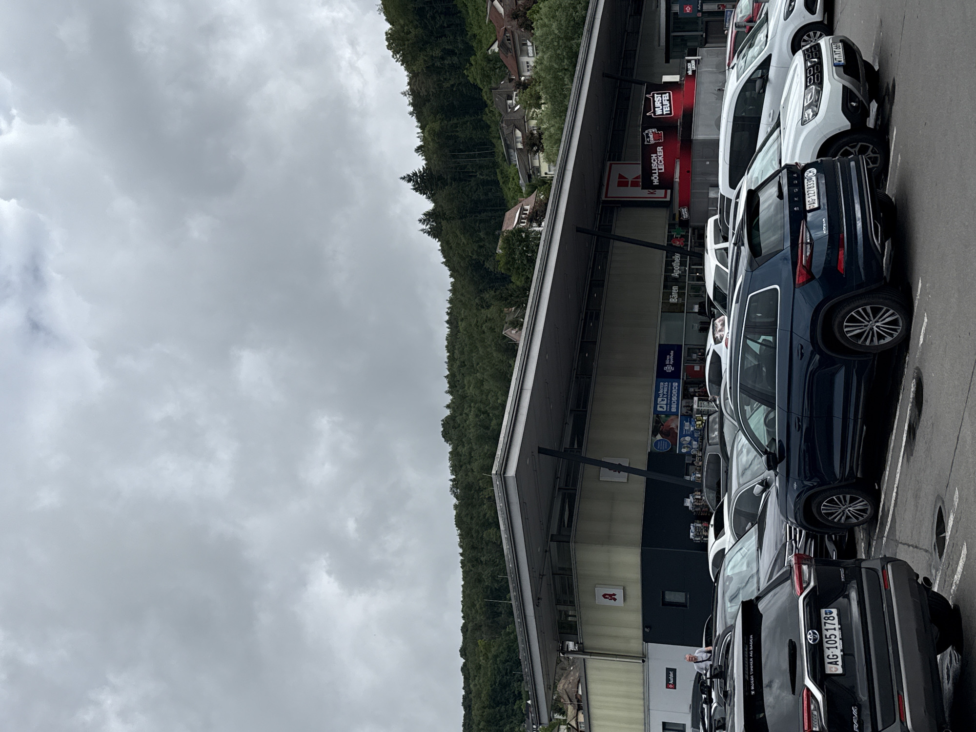 The image displays an outdoor scene of a commercial parking lot with a multi-story building alongside it, backed by a lush, forested hill under an overcast sky. Several cars are parked in designated spots.
In the foreground, a black Toyota RAV4 Hybrid with a Swiss license plate "AG-105178" is prominently visible. Next to it is a dark blue Skoda SUV with the Swiss plate "AG-127833 CH". Further down the row, a white Dacia Duster bears the German license plate "WTXT 669". Other vehicles are partially visible, including a white van and another SUV. One person can be seen near the black Toyota RAV4, appearing to be walking or standing.
The adjacent building features various storefronts and signs. Prominently displayed are signs for "WURST TEUFEL" (Sausage Devil) and "HÖLLISCH LECKER" (Hellishly Delicious), suggesting a food establishment. A large "K" logo is also visible, often associated with a German supermarket chain. Other visible texts include "Bären Apothek", "Apothek", "Raxon", "Audifahrt", "Mister Press", and "RUEDI TINNER AG BADEN".
The scene suggests a typical daytime shopping or commercial area, likely located near the German-Swiss border given the mix of German (WT for Waldshut-Tiengen) and Swiss (AG for Aargau) license plates, consistent with the provided context of Waldshut, Germany. The sky is completely covered with thick grey clouds, indicating an overcast or potentially rainy day.