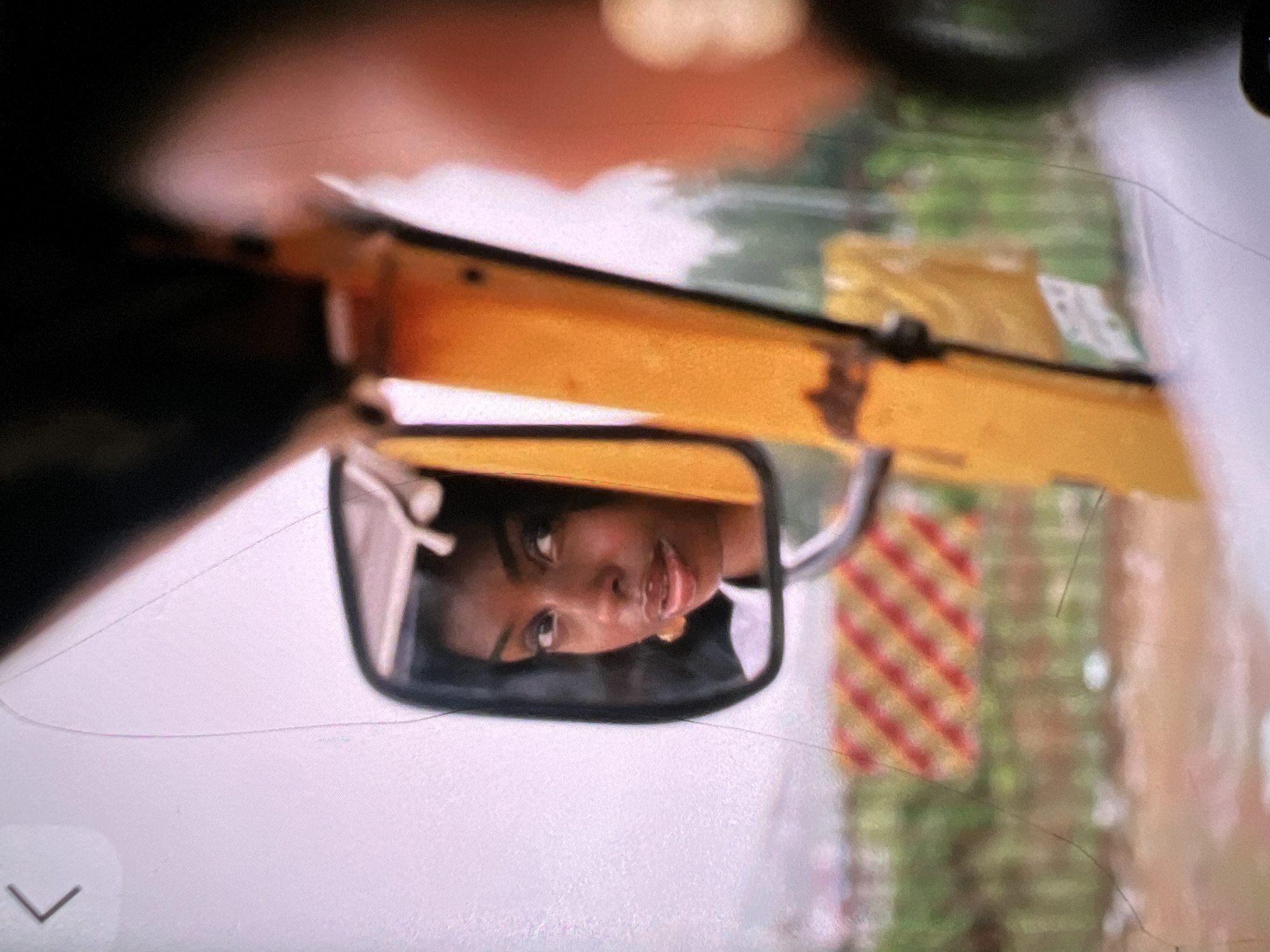 The image captures a young Black woman looking into a side-view mirror, her reflection is seen in the mirror. The woman is wearing a white top and a gold earring. She has dark hair and her lips are slightly parted. The mirror itself is mounted on a yellow structure, which appears to be part of a vehicle, possibly a commercial bus or a truck. The background shows a street scene with a blurred checkered pattern of red and yellow in the distance, suggesting a roadside barrier or advertisement. Green foliage and a muddy road surface are also visible, indicative of an urban or semi-urban environment. The lighting suggests an overcast or indoor setting. The overall impression is a candid moment captured while on the move, likely within the city of Lagos, Nigeria, given the visual cues of a bustling environment and common vehicle types.