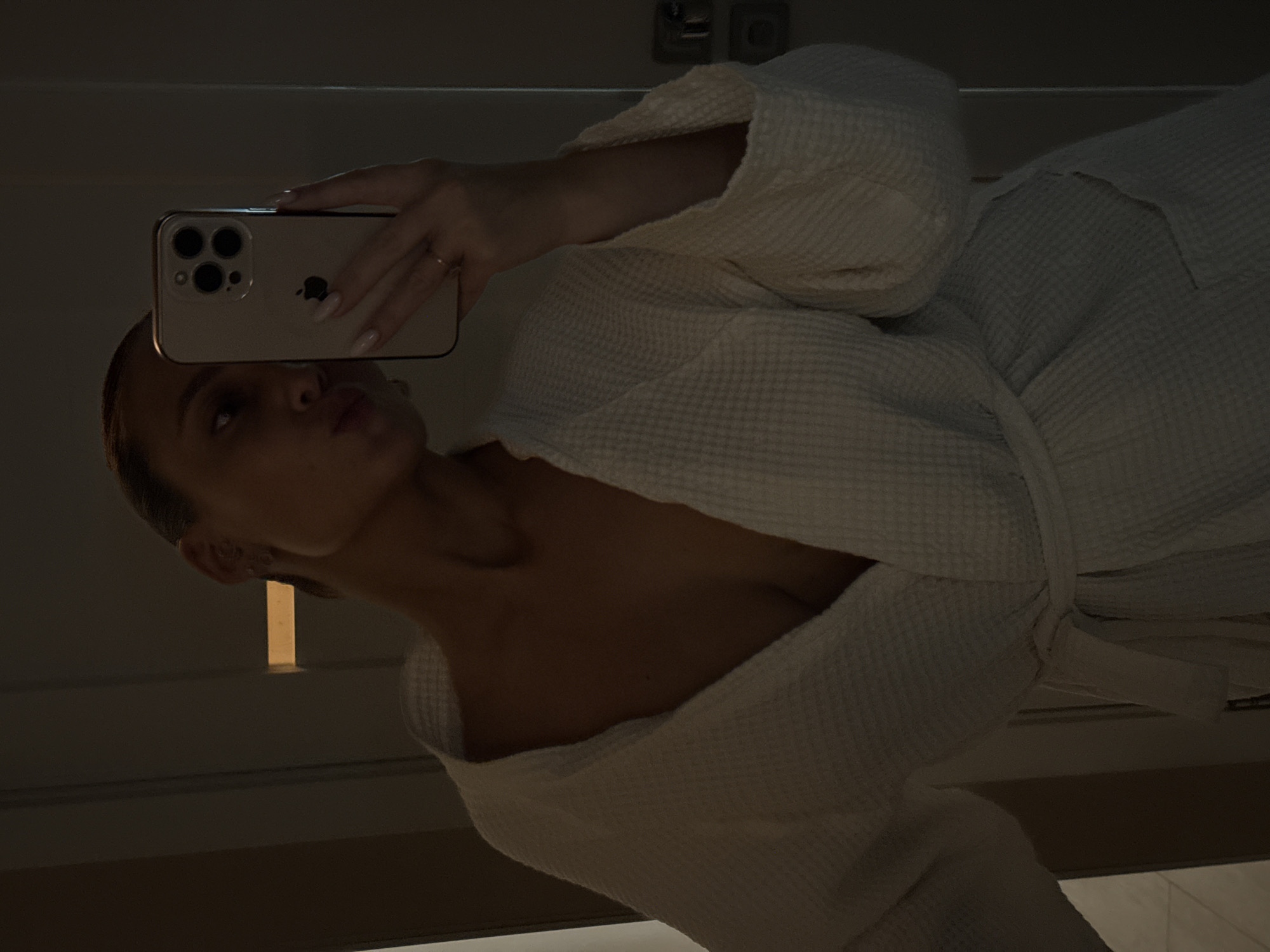 A woman is taking a selfie in a mirror. She is wearing a white bathrobe and holding a gold iPhone with three rear cameras. The environment is dimly lit, and the location is unspecified in terms of Chaikovsky, Russia.
