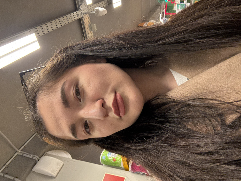 A young adult female is depicted in a close-up, chest-up shot within a retail store environment located in Kurovskoye, Russia. She has long, dark brown hair, parted slightly off-center, cascading over her shoulders. Her face is presented frontally, with light brown eyes gazing directly at the viewer. She exhibits a neutral expression with a slight pout, wearing a muted pink or nude lipstick. Her attire consists of a light beige or tan woolen-texture coat worn over a white, ribbed long-sleeved top, partially visible at the neckline.
The background, largely out of focus, reveals elements of a commercial interior. To the viewer's left, stacks of wrapped consumer goods, including items with green and pink packaging, are partially visible. A portion of a red sign or fixture is also present. To the viewer's right and above, the ceiling features exposed utilities: a network of silver-colored cable trays or conduits, rectangular fluorescent light fixtures providing artificial illumination, and a white, dome-shaped security camera or sensor mounted on the ceiling. The overall lighting is bright and even, consistent with an indoor retail space.