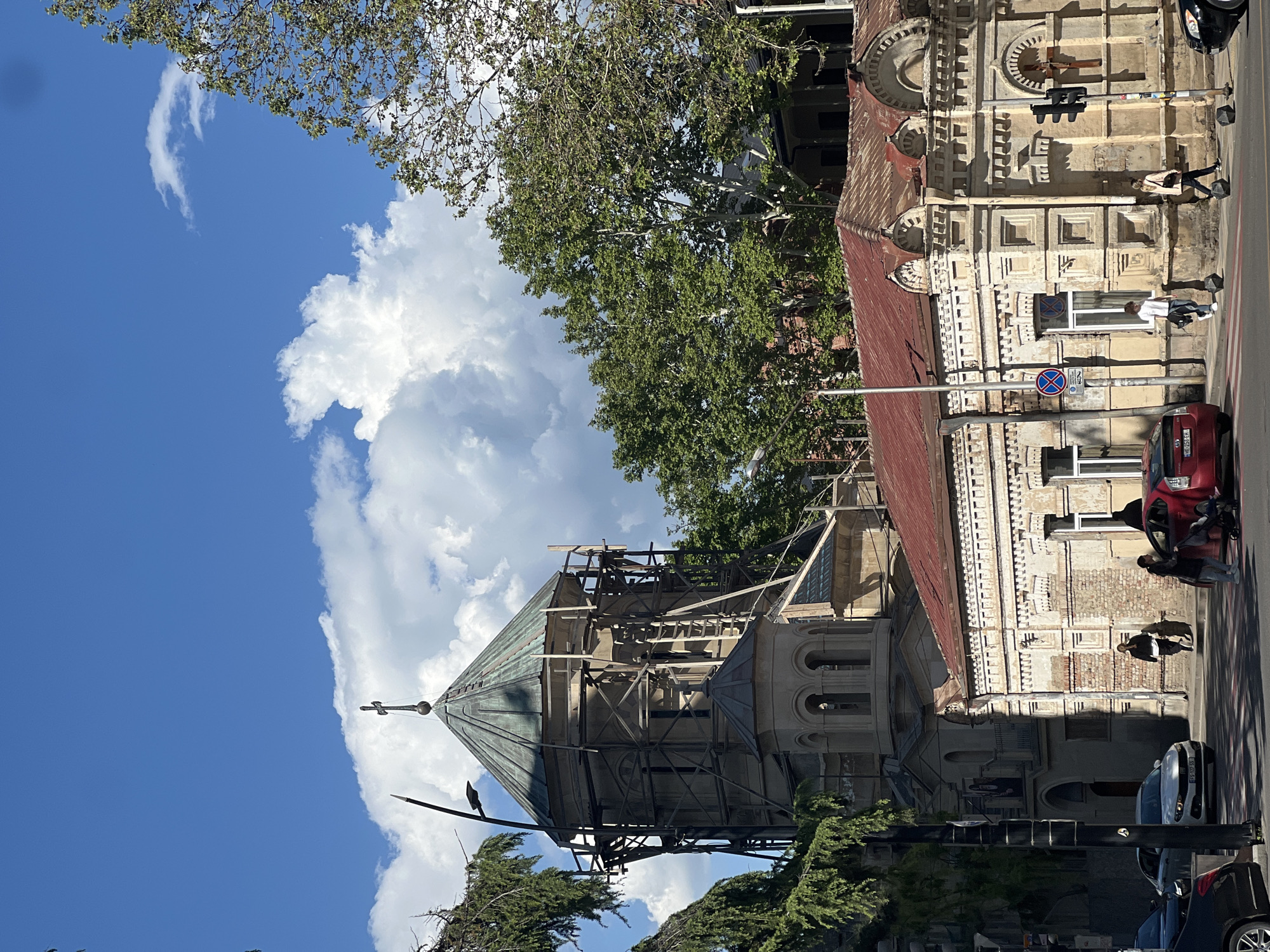 The image shows a street scene in Tbilisi, Georgia. A tall building, possibly a church, is under renovation with scaffolding surrounding its conical roof. The building has detailed architectural features. A red-roofed structure adjoins the church.
On the sidewalk, two pedestrians walk past a light-colored building with a red roof. Several parked cars are visible along the street. Above, a clear blue sky is filled with large, puffy white clouds. A few trees with green leaves frame the buildings.