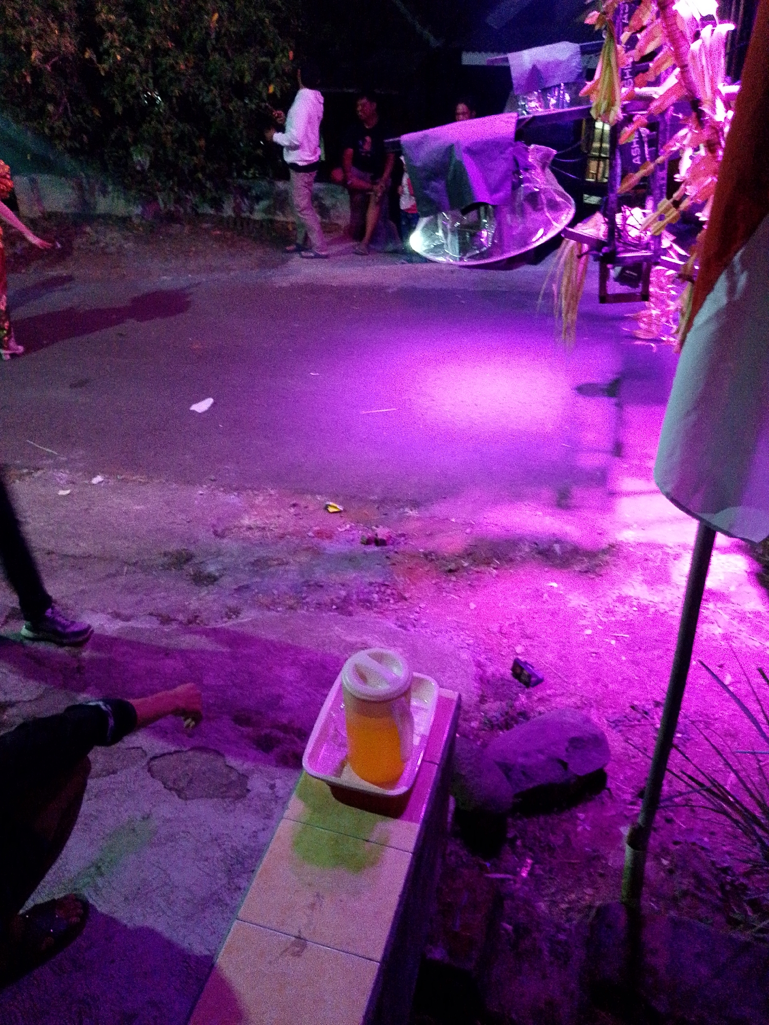 In Babadan, Indonesia, an outdoor scene unfolds under the vibrant glow of purple lighting. A street or open paved area is visible, with some dirt and debris scattered on the ground. To the left, a person in dark clothing is partially visible, with their arm extended. In the foreground, a white tray holds a translucent plastic cup filled with a yellow liquid, topped with a white lid. Beside the tray, a large, dark-colored rock rests on the ground. A slender, dark pole, likely a bamboo stalk, stands vertically on the right side of the frame, with some greenery visible near its base. In the background, illuminated by the purple lights, is what appears to be a sound system or stage setup, with decorative elements that resemble dried corn husks. Several individuals are present, including one person in a white shirt and light-colored pants standing near the background setup, and another in a dark shirt and shorts to their right. Further left, a person in a red garment with patterned details is seen from the side, with their left arm extended. The overall atmosphere suggests a gathering or event taking place at night.