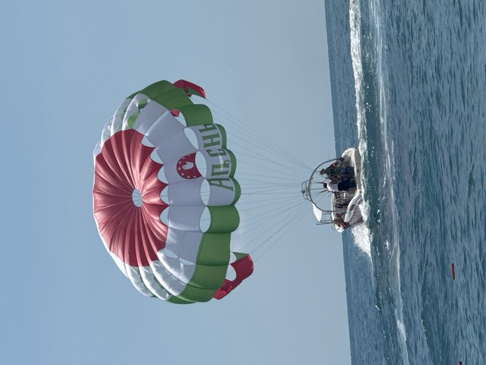 In Gagra, Abkhazia, a parasail with a red, white, and green design, featuring Cyrillic lettering and a hand symbol, is suspended against a clear blue sky. The parasail is tethered to a white motorboat that is moving through choppy blue water, creating a white wake. Several people are visible on the boat, some appearing to be passengers and others potentially operating the vessel or supervising the parasailing activity. The overall scene depicts a recreational activity taking place on the sea.