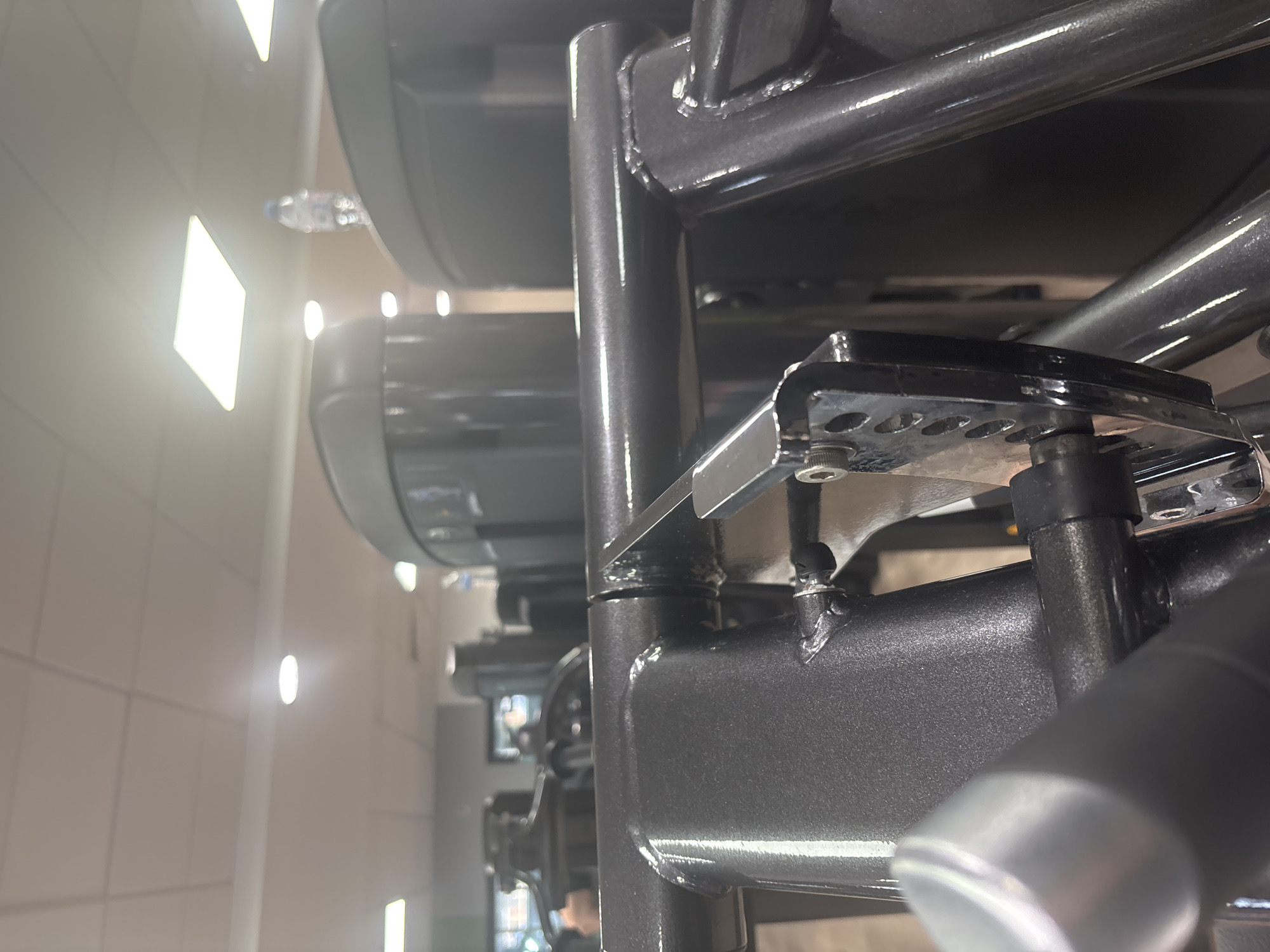 The image depicts a close-up view of a weight rack in a gym. The rack is made of gray metal and has multiple levels for holding dumbbells. The lighting in the gym is bright, with fluorescent lights visible in the background. In the background, a water bottle can be seen behind the rack. The image focuses on the structural details of the rack, including the welds and bolts that hold it together. The gym setting suggests an indoor environment dedicated to physical fitness. There is no indication that this image was taken in London, United Kingdom.