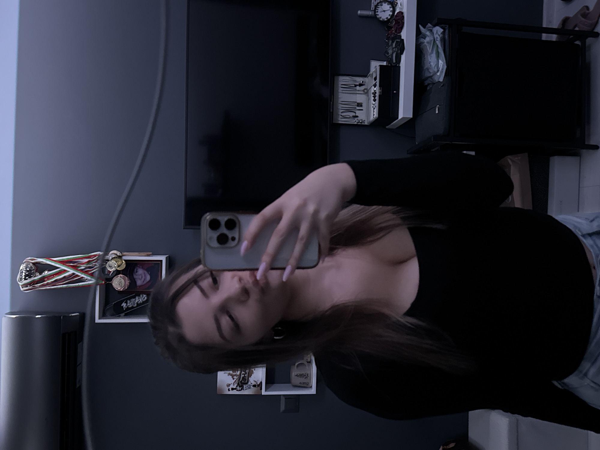 A young woman is taking a selfie in what appears to be an apartment or living space. She's wearing a black long-sleeved top and light blue denim jeans and is holding a smartphone, presumably to take the photo. The environment includes a television mounted on the wall, a white shelving unit with decorative items, possibly medals and a framed picture hanging on the wall to the left, and a white sofa in the foreground. There is no information available from the image that could confirm a location in Sofia, Bulgaria.