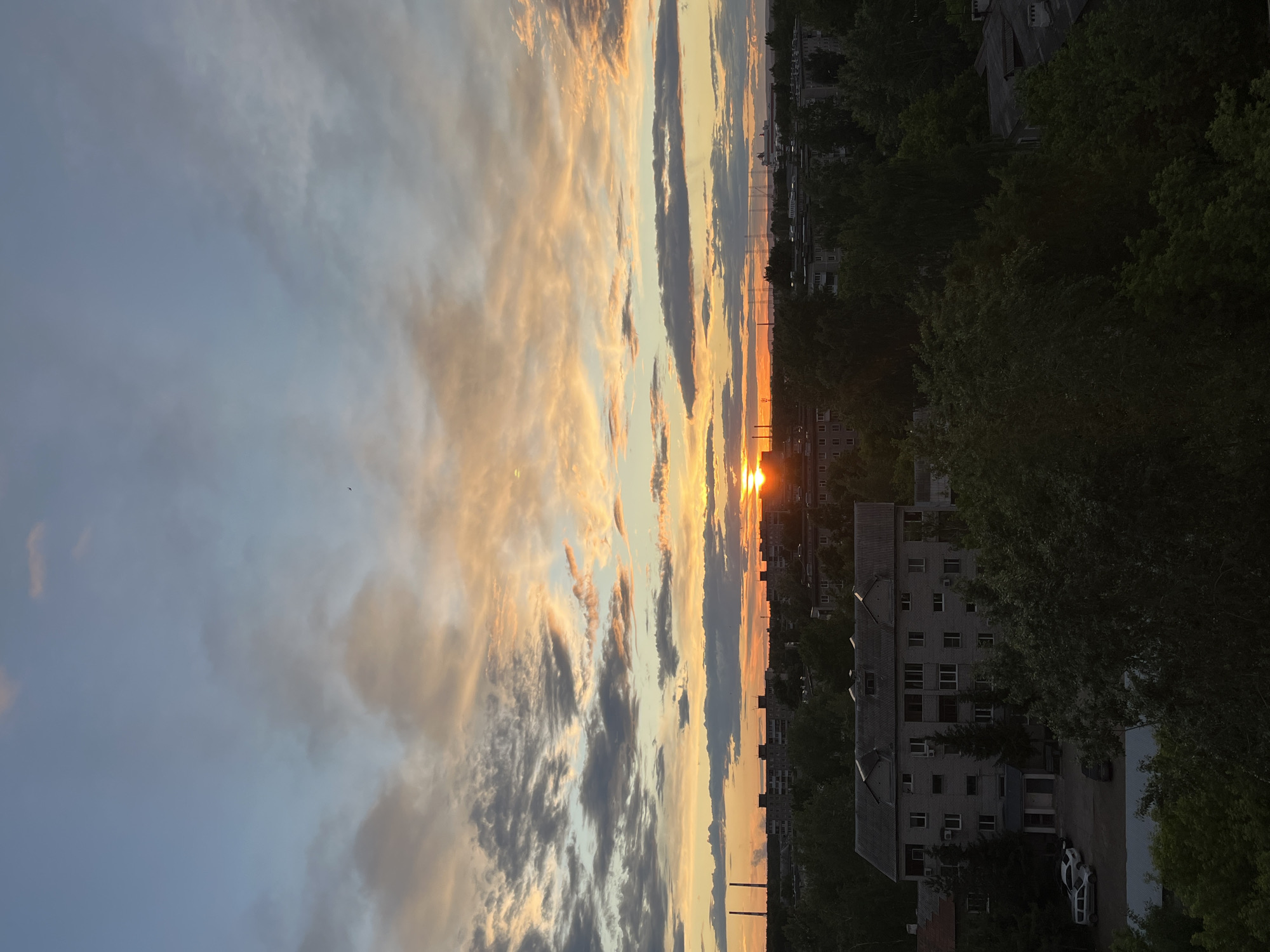 The image depicts a sunset over a cityscape. In the sky, various cloud formations are visible, tinged with orange and yellow hues from the setting sun. The sun itself is a bright, distinct point on the horizon. Below the sky, there are apartment buildings and trees silhouetted against the fading light. A white car is parked next to a building on the street. The image appears to be taken from a high vantage point. The location is Unknown, Unknown.