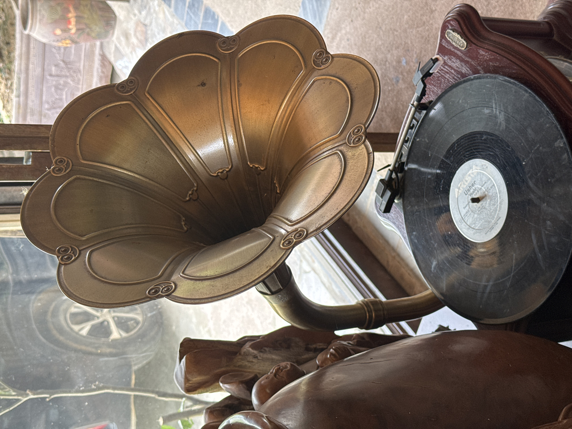 This detailed image captures a vintage brass gramophone, centered prominently in the foreground, within an indoor setting that overlooks an outdoor scene in what is understood to be Bản Đêu, Vietnam.
The primary subject is a classic phonograph featuring a large, ornate, multi-lobed horn crafted from a warm, aged brass or bronze-colored metal. The horn exhibits decorative raised patterns, including curved ridges and stylized circular motifs resembling coiled eyes at the scalloped edges. It connects via a curved, brass-colored arm to a dark, reddish-brown wooden base with a polished finish and an antique aesthetic, which includes a small, rectangular brass-colored plaque on its right side. A black vinyl record with a light-colored center label, partially legible as "Usher" and "Yeah!", rests on the turntable platter. A black tonearm with a cartridge is positioned above the record.
To the immediate left of the gramophone, a section of a dark brown, intricately carved wooden sculpture or decorative object is visible, displaying rounded forms and indistinct human-like figures or faces.
The background reveals an outdoor environment seen through a clear window. A reflective surface on the left side of the window shows a clear reflection of a dark-colored vehicle, specifically a tire with a multi-spoke alloy wheel, suggesting a street or parking area nearby. A thin tree branch with sparse foliage is also visible against the windowpane. On the right side of the outdoor view, two large ceramic planters are present: one lighter-toned with a detailed bas-relief pattern, and another taller, reddish-brown pot adorned with painted red and green lotus flowers. The ground directly outside the window appears to be a light-colored tiled patio or walkway. The scene is illuminated by natural light entering through the window, casting highlights and reflections on the gramophone's metallic surfaces. There are no people visible in the image.