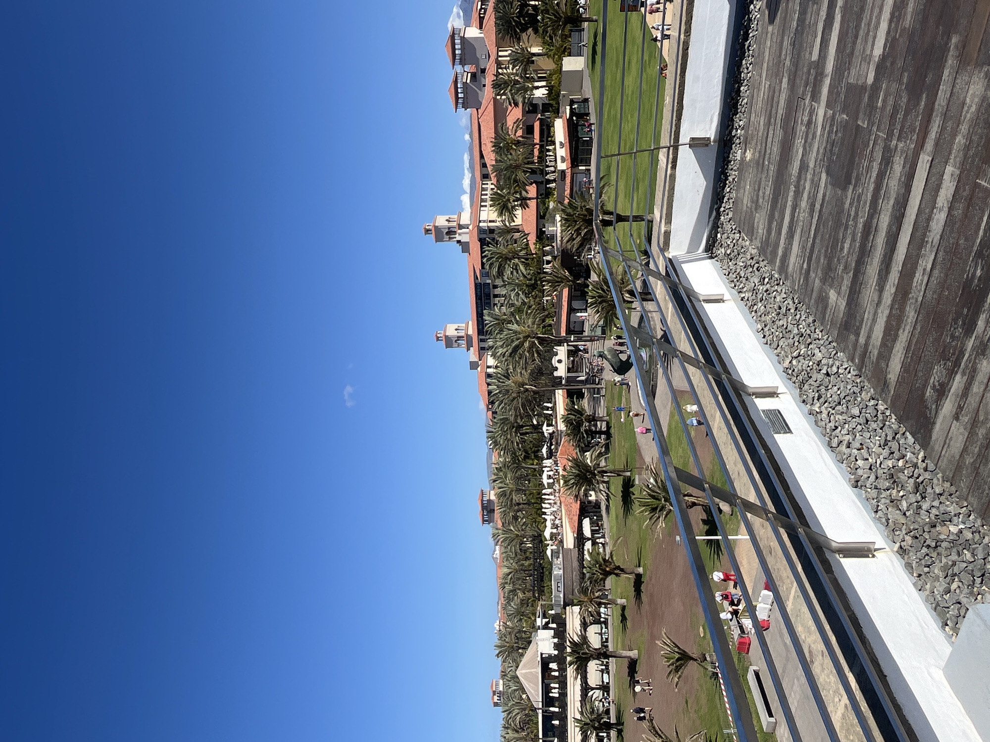 The image shows a view from an elevated position in Maspalomas, Spain, looking towards a resort complex with multiple buildings featuring red tile roofs and palm trees. Below the buildings, there are well-maintained green lawns, walkways, and recreational areas with visible people and activities. A silver railing and a deck area are in the foreground, suggesting the viewpoint is from a balcony or higher floor of a building. The sky is clear blue. The overall scene depicts a sunny, outdoor resort environment.