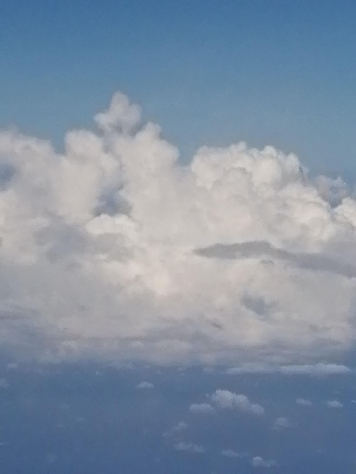 The image shows a view of clouds in a blue sky. The clouds are predominantly white and gray, with varying densities and shapes. The sky is a lighter blue at the top, transitioning to a darker blue at the bottom of the frame. The image location is Unknown, Unknown.