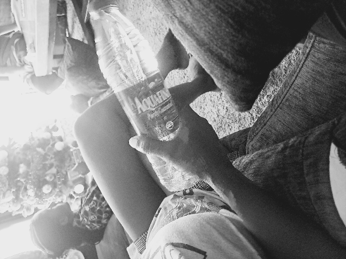 The monochrome image depicts an indoor scene in Ikate, Nigeria. In the immediate foreground, a person's right arm and hand are visible, holding a clear plastic bottle labeled "AQUAFINA." This person wears a light-colored short-sleeved top and blue denim jeans. A bare foot is partially visible on the carpeted floor. In the midground, another individual is reclining, with their bare legs prominently positioned. This person is dressed in patterned shorts and a top, with their head visible in profile, facing left towards a brightly lit area. Behind this individual, a decorated Christmas tree adorned with ornaments and lights is prominently displayed. In the background, elements of furniture and what appears to be another person's lower legs are faintly visible, illuminated by strong ambient light. The floor throughout the visible area is carpeted.