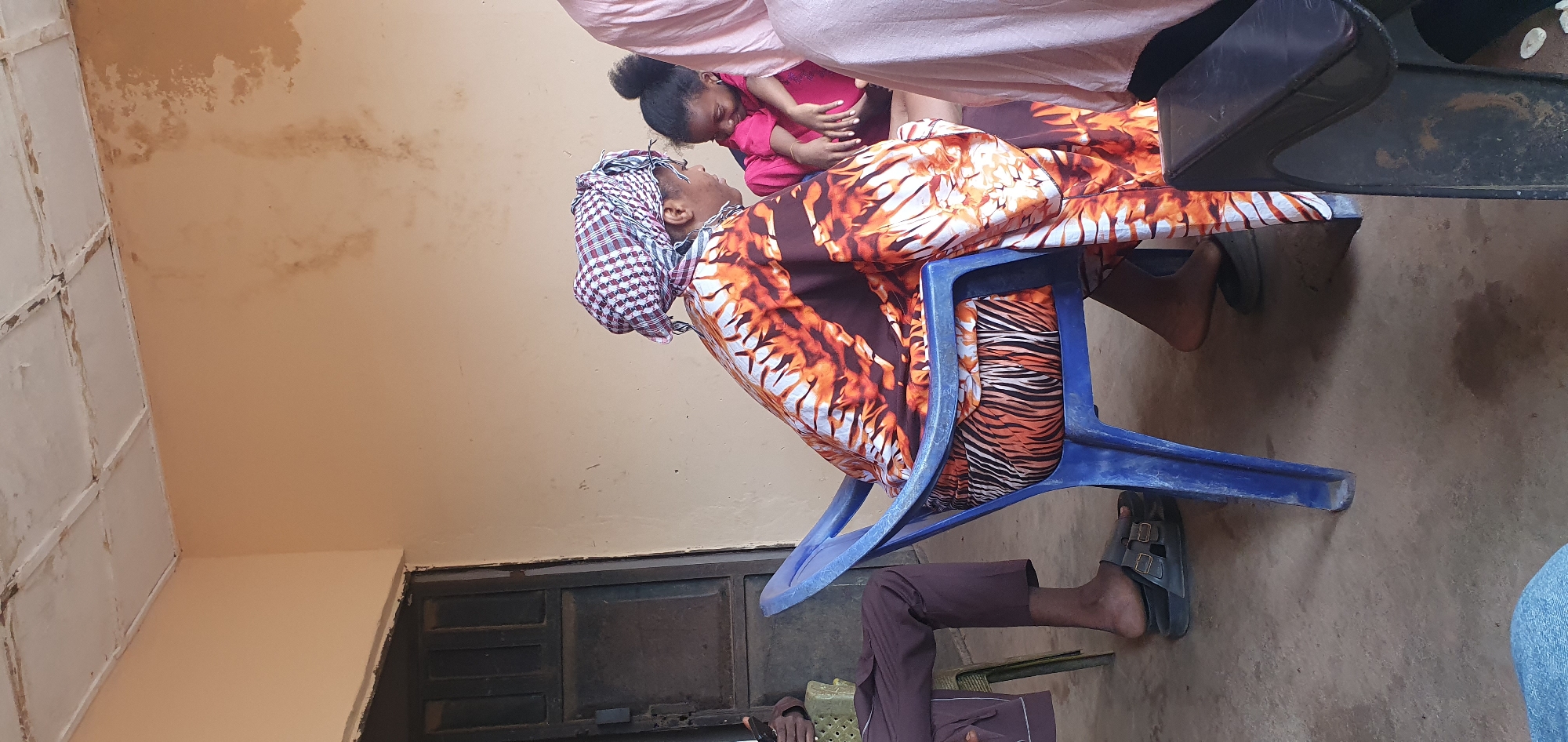 The image depicts an intimate interaction between an adult and a small child within an indoor or sheltered setting in Idah, Nigeria. An adult is seated on a blue plastic chair, wearing a vibrant orange, black, and white patterned garment, possibly a dress or wrapper, and a checkered headwrap. Their face is visible in profile, looking up at a young child in a pink top, who is leaning into or being held by the adult. The child's profile is also visible, looking down at the adult, suggesting a tender and affectionate moment.
The background features light brown or tan walls with noticeable areas of discoloration and water stains, indicative of an older building or maintenance issues. To the upper left, a white ceiling or panel with a grid-like pattern is visible. A dark, possibly metal, door or cabinet structure can be seen on the lower left. The floor is a light, neutral-toned concrete or dirt surface. Partially visible in the foreground are the legs and feet of another person, wearing dark pants and open-toed sandals, seated on a chair next to the main subjects. The lighting is diffused, suggesting a daytime scene. No visible text is present in the image. The scene conveys a sense of familial warmth in a modest local environment.