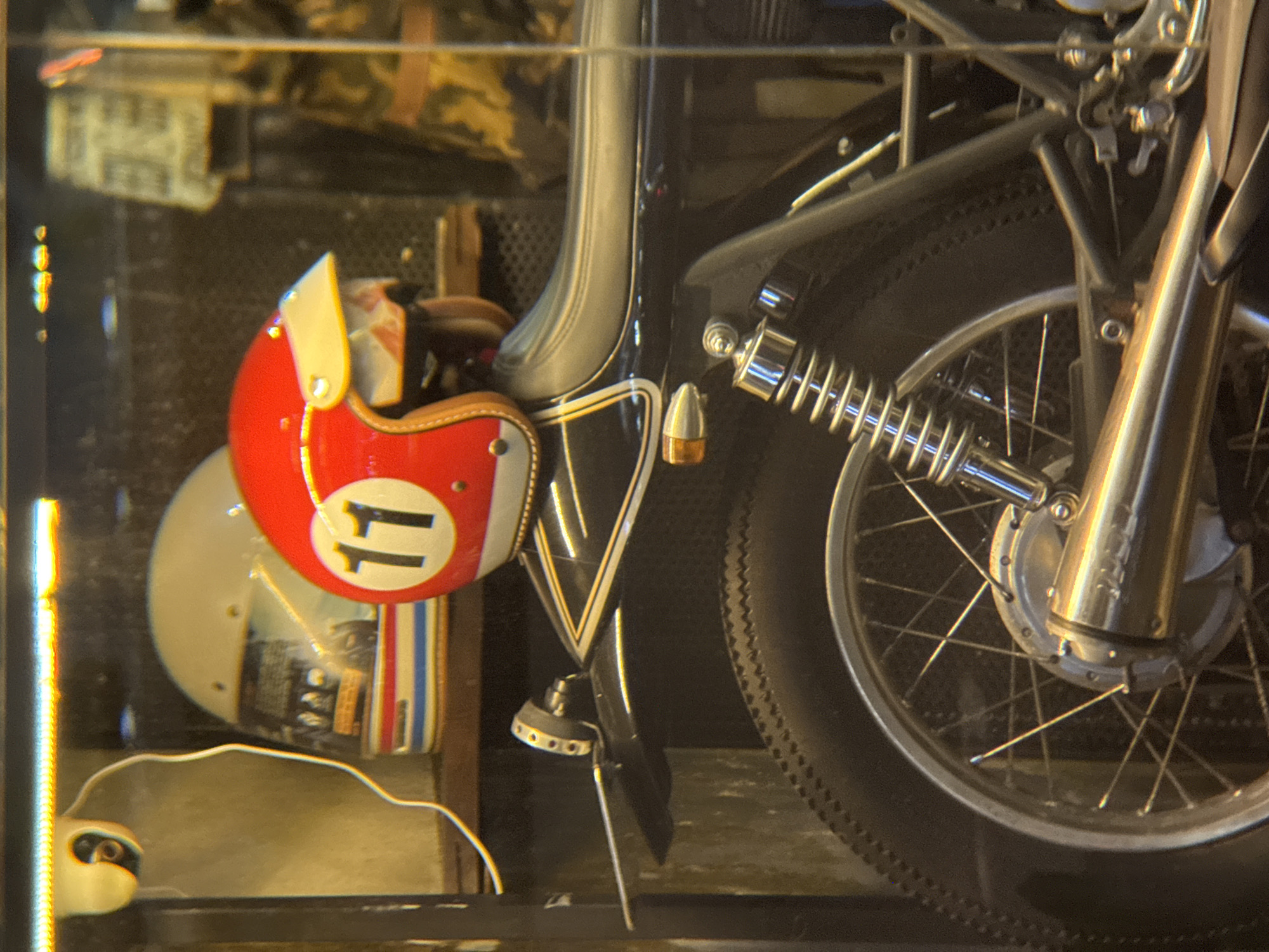 This is a close-up, eye-level, medium shot of a vintage motorcycle parked indoors. The motorcycle is black and partially visible from the rear wheel up to the seat. The rear wheel is a spoked wheel with a black tire. A chrome shock absorber is visible behind the wheel. The motorcycle seat is black and appears to be made of leather.
Above and behind the motorcycle, there are two motorcycle helmets. The helmet on the left is white with red, white, and blue stripes around the base and has a reflective visor. The helmet on the right is predominantly red with a white circle on the side containing the black number "11". This helmet also has a yellow visor. A white wire extends from the upper left corner of the frame, connected to a small, white, dome-shaped object, possibly a security camera. The background is blurred and appears to be a retail or display space with shelves and some items on them. The overall lighting is warm and somewhat dim, suggesting an indoor setting possibly in a shop or garage.
The image is taken in Thuận Hóa Ward, Vietnam.