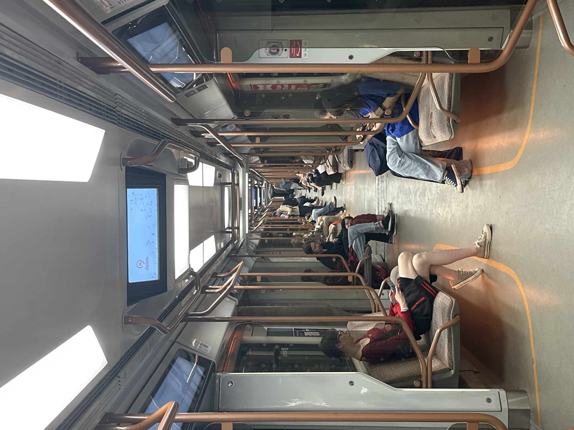 The image depicts the interior of a Moscow Metro train car, likely during a non-peak hour, with several passengers seated and standing. The train car features light-colored walls and ceiling with prominent fluorescent lighting fixtures. Copper-colored poles and handrails are visible throughout the car, providing support for passengers. Rows of seats are arranged on either side of the aisle, upholstered in a patterned fabric.
In the foreground on the left, a woman with dark hair, wearing glasses, a red top, and a black skirt, is seated and looking at her phone. To her right, another passenger is partially visible. On the right side of the aisle, a woman with long dark hair, wearing a blue top and light-colored jeans, is seated and also looking at her phone. Another passenger is seated next to her, mostly obscured. Further down the car, more passengers are seated, some with their backs to the camera. A digital screen is mounted overhead, displaying a map or information related to the metro system. The floor of the train car is a light gray color with a bright yellow line marking the edge of the platform or a safety zone. The overall impression is one of a functional, modern public transportation environment.
