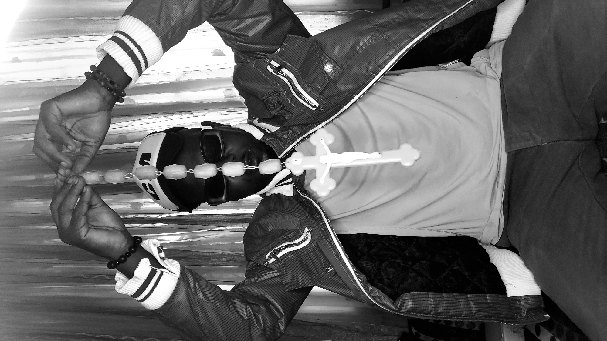 A black and white, medium shot photograph shows a person sitting, holding a rosary with a crucifix pendant. The person is wearing a jacket with white and black striped cuffs, sunglasses, and a baseball cap. The background consists of a curtained window. The image is a selfie or taken by someone close to the subject, as indicated by the close-up framing and the subject's direct engagement with the camera. The location of Sabon Tasha, Nigeria, is not visually indicated in the image itself.