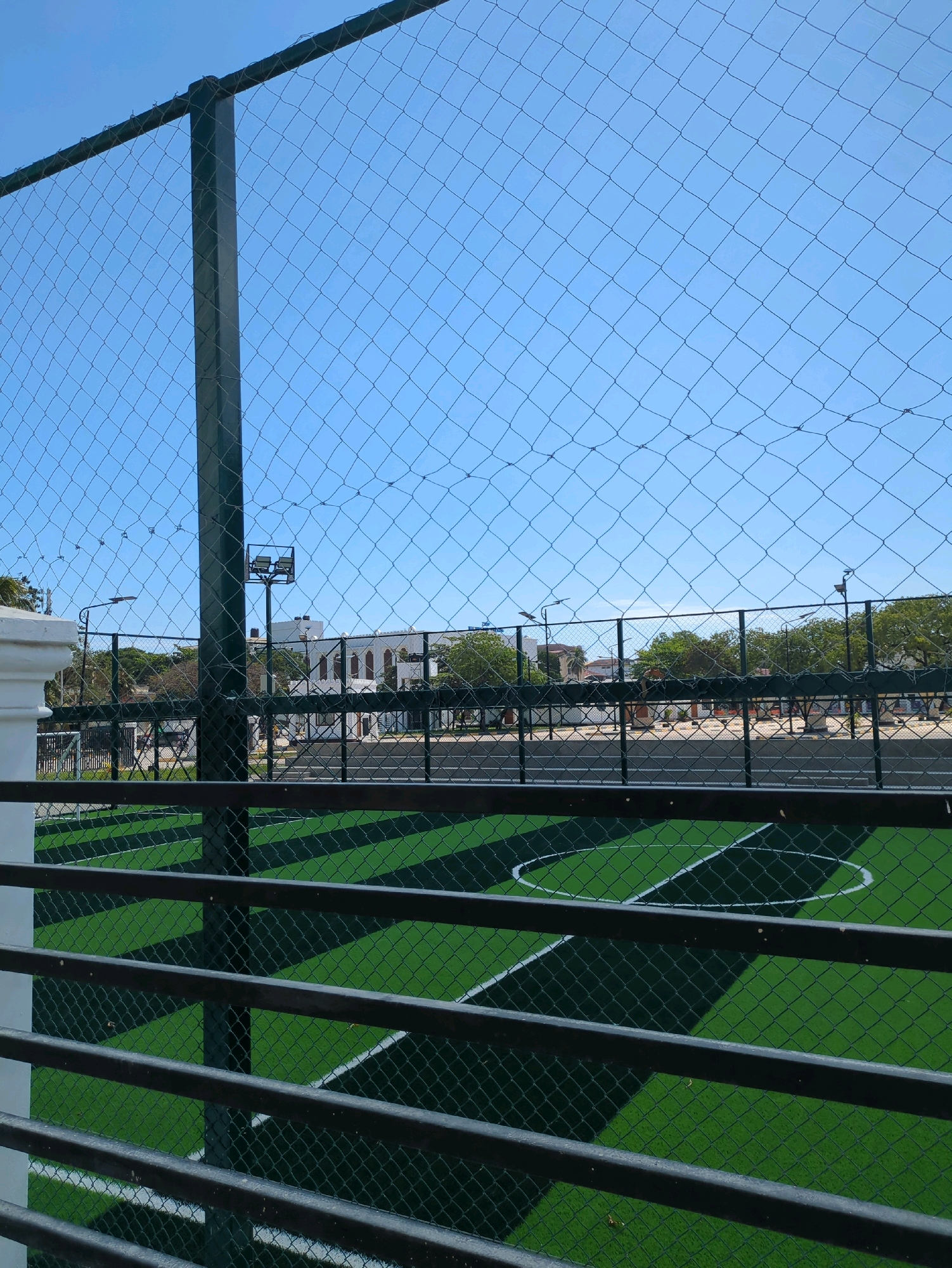 The image captures a view of an artificial turf football pitch through a metal fence and netting. The pitch is marked with white lines, indicating its boundaries and a circular area. In the background, there are buildings with architectural features characteristic of Zanzibar, including arched windows and possibly coral stone construction. Trees and streetlights are also visible, suggesting an urban or semi-urban environment. The sky is clear and blue. The overall scene depicts a recreational facility, likely a sports complex or a school ground, situated in Zanzibar, Tanzania.